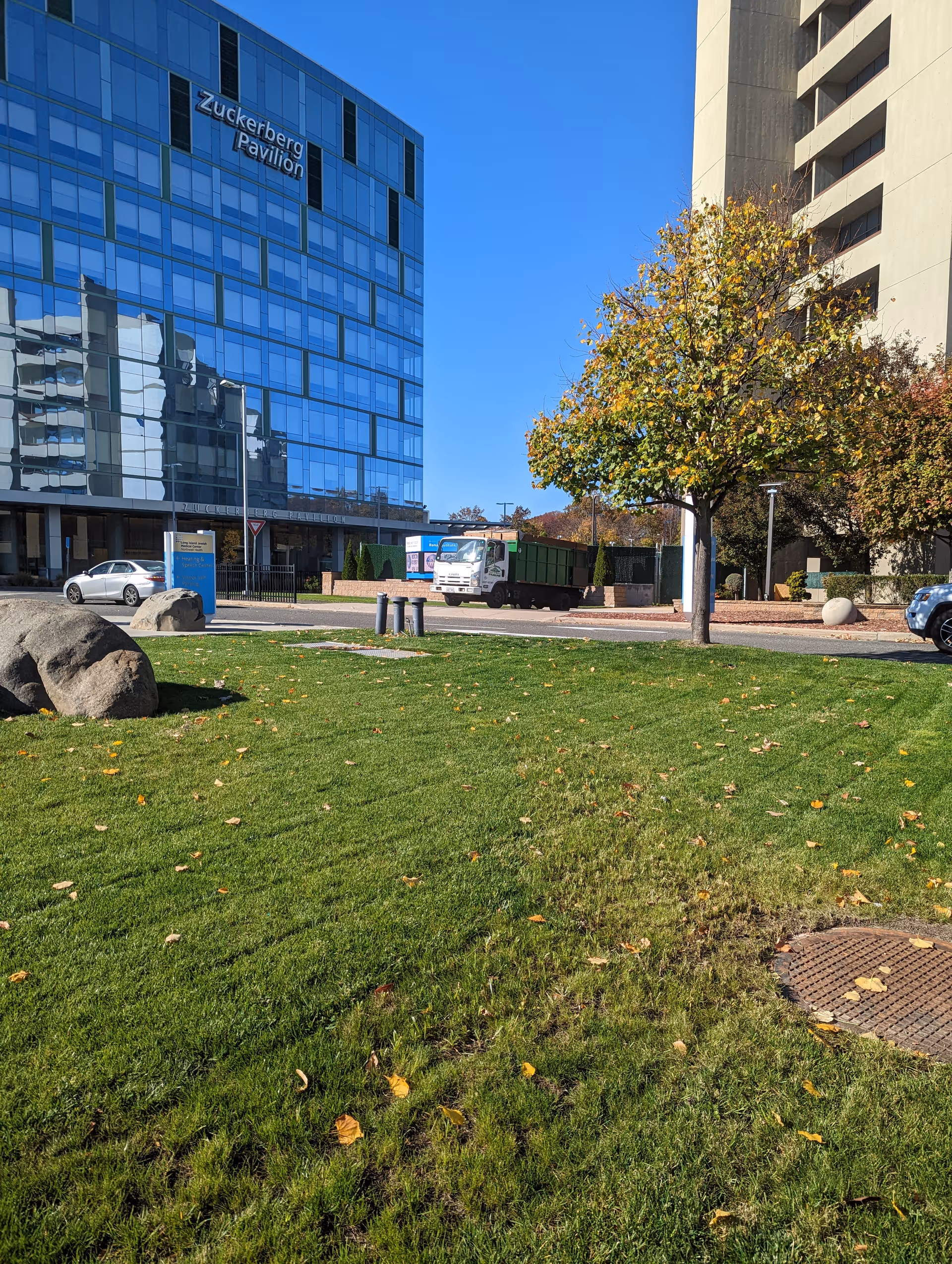Modern glass-faced building labeled Zuckerberg Pavilion with grassy foreground, tree, and street vehicles.
