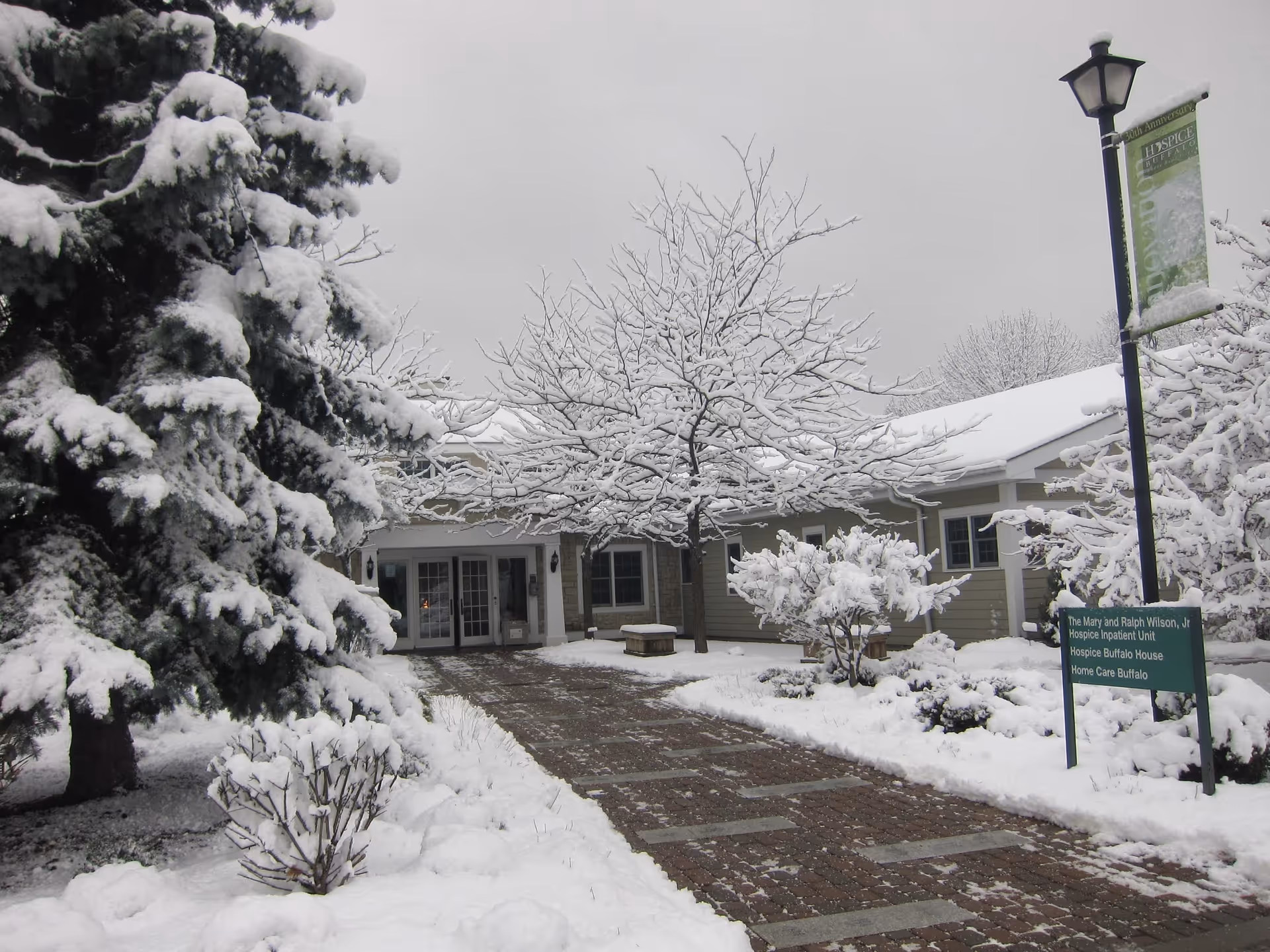 Snow covers the front of a hospice building, with trees, a sign, and a brick walkway.