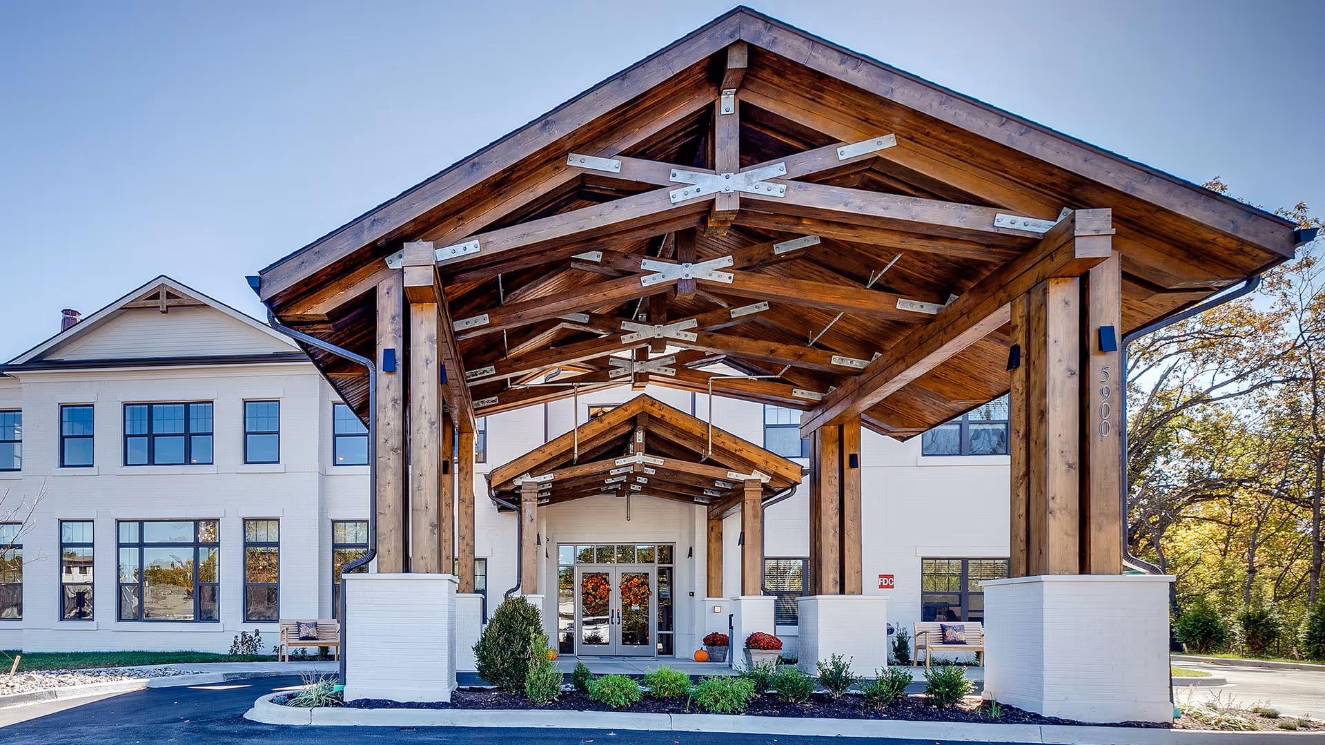 Front exterior view of Everlan of Louisville facility showing a large covered entrance with wooden beams and white brick pillars, landscaped greenery, and glass double doors decorated with autumn wreaths.