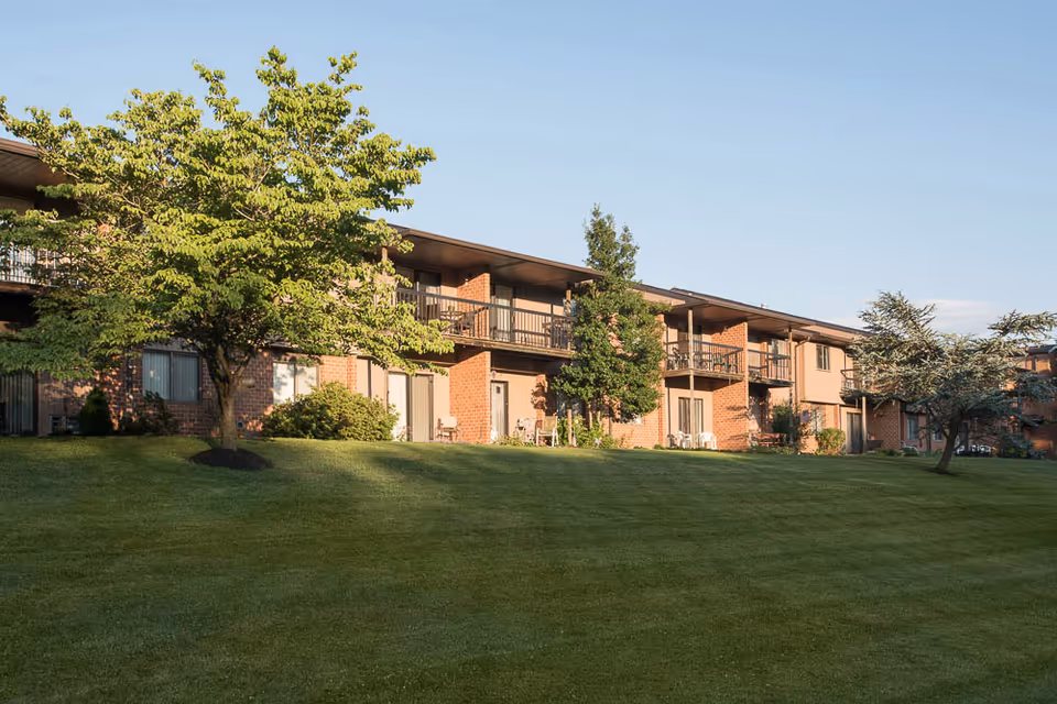 Exterior view of a two-story brick retirement community building with balconies and patios, surrounded by green grass and several trees under a clear blue sky.