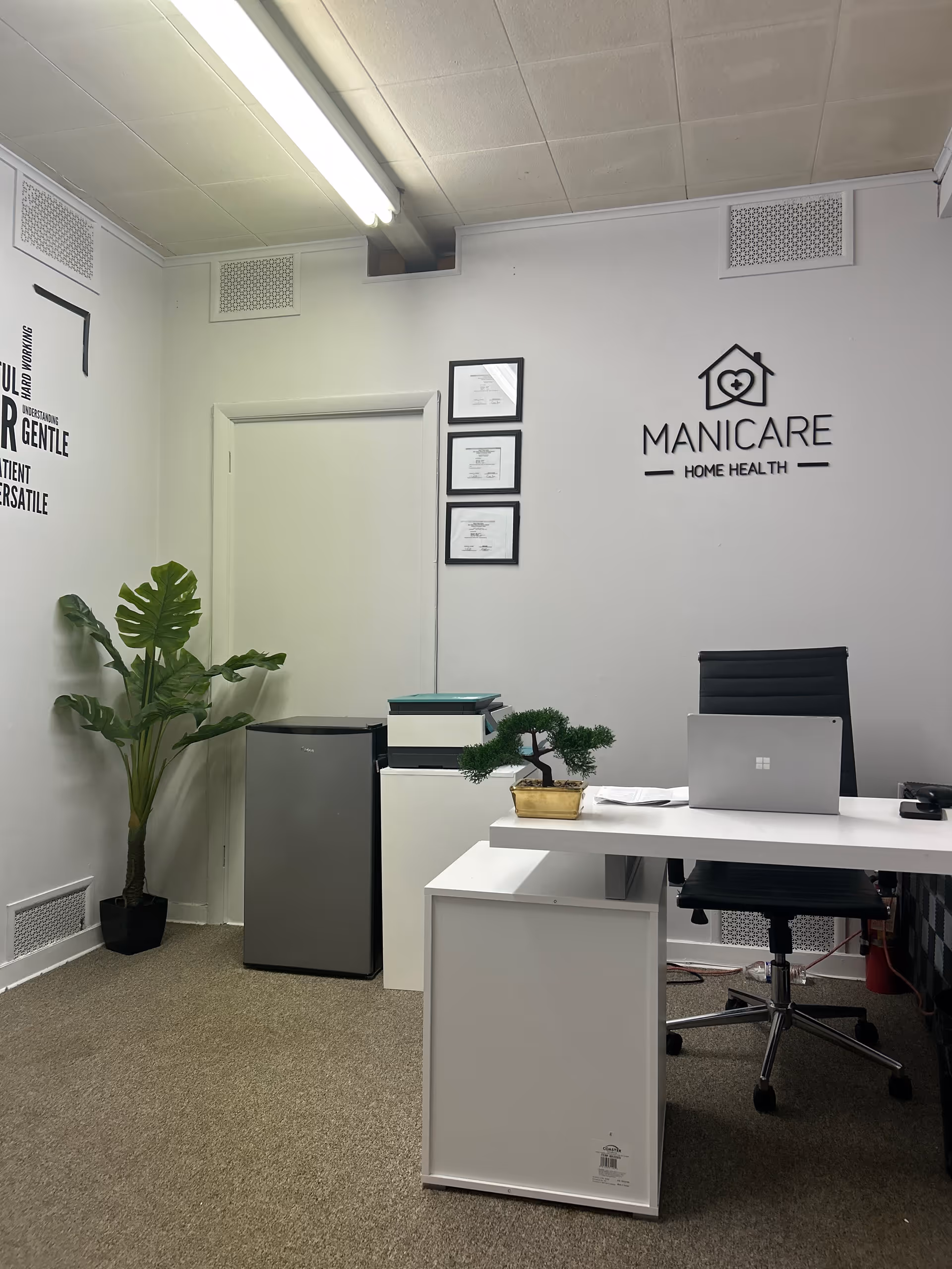 Office reception area with Manicare Home Health logo, framed certificates, desk, laptop, and plant.