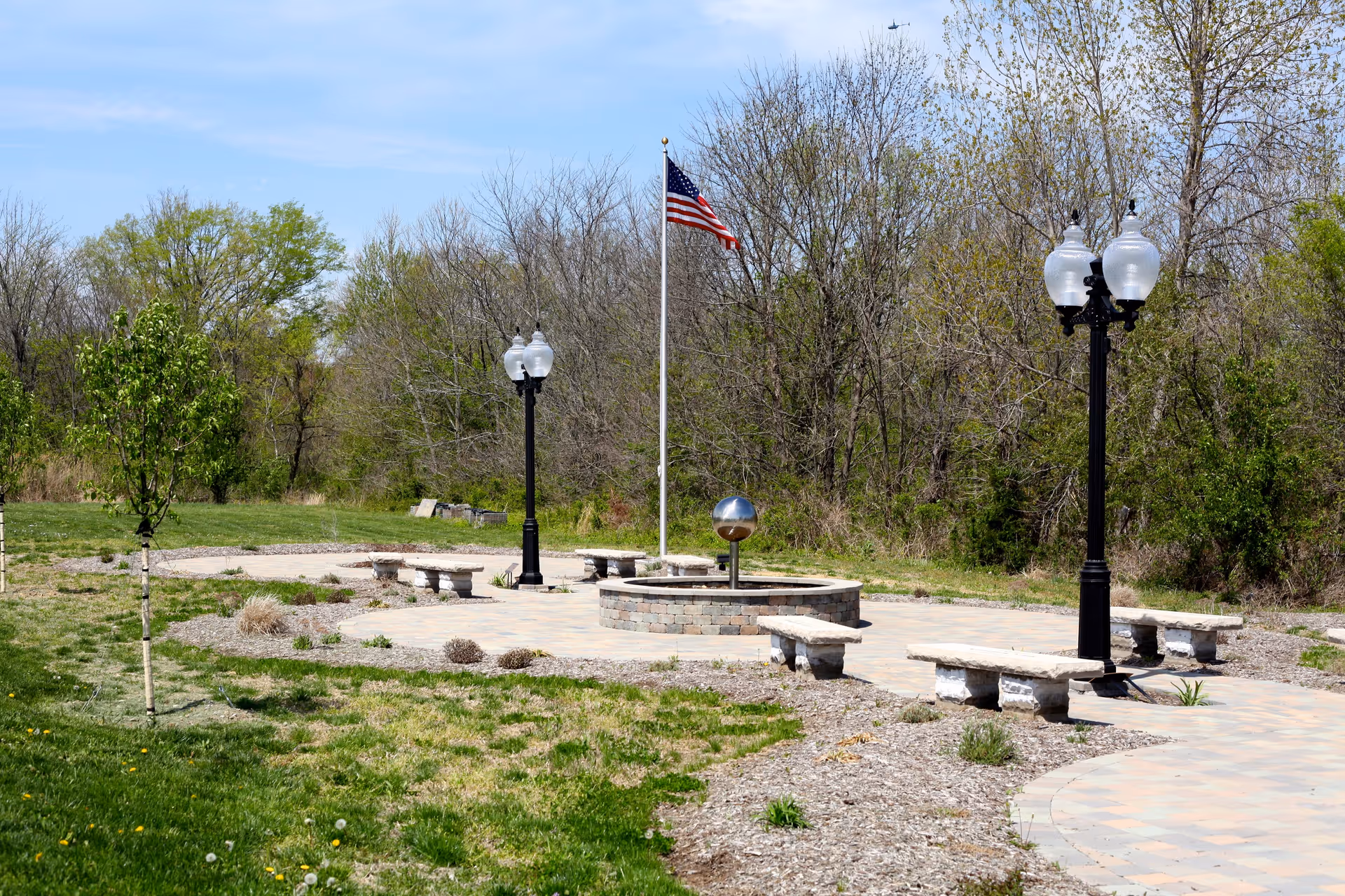 Outdoor memorial plaza with flagpole, streetlamps, circular seating, and surrounding trees.