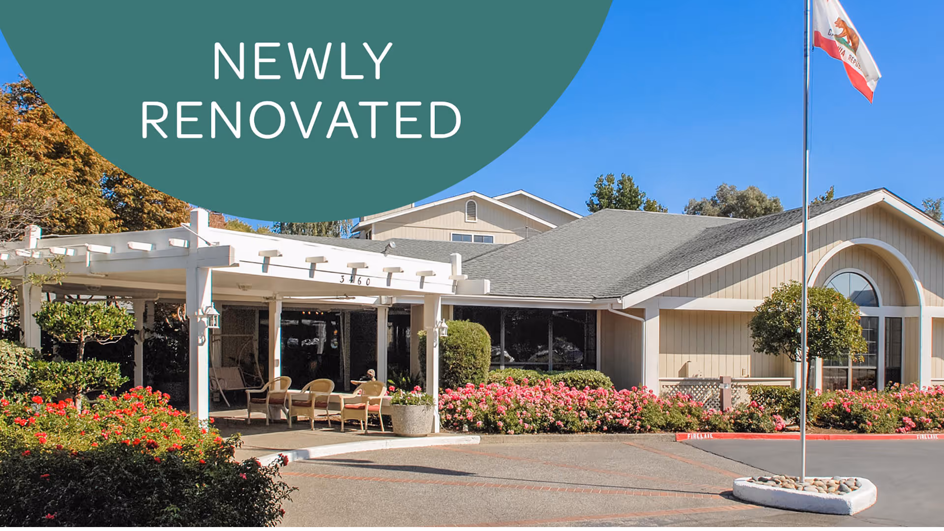Exterior view of a newly renovated senior living facility building with a covered entrance, outdoor seating, and well-maintained landscaping including bushes and flowers. A flagpole with a California state flag is visible in front of the building under a clear blue sky.