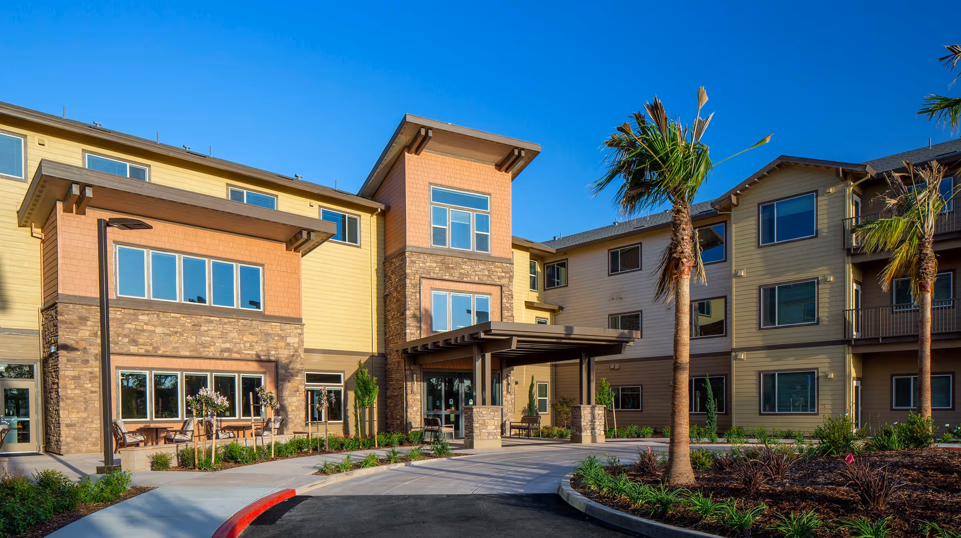 Exterior view of Bruceville Point senior living facility showing a modern three-story building with stone and beige siding, large windows, a covered entrance, palm trees, and landscaped grounds under a clear blue sky.