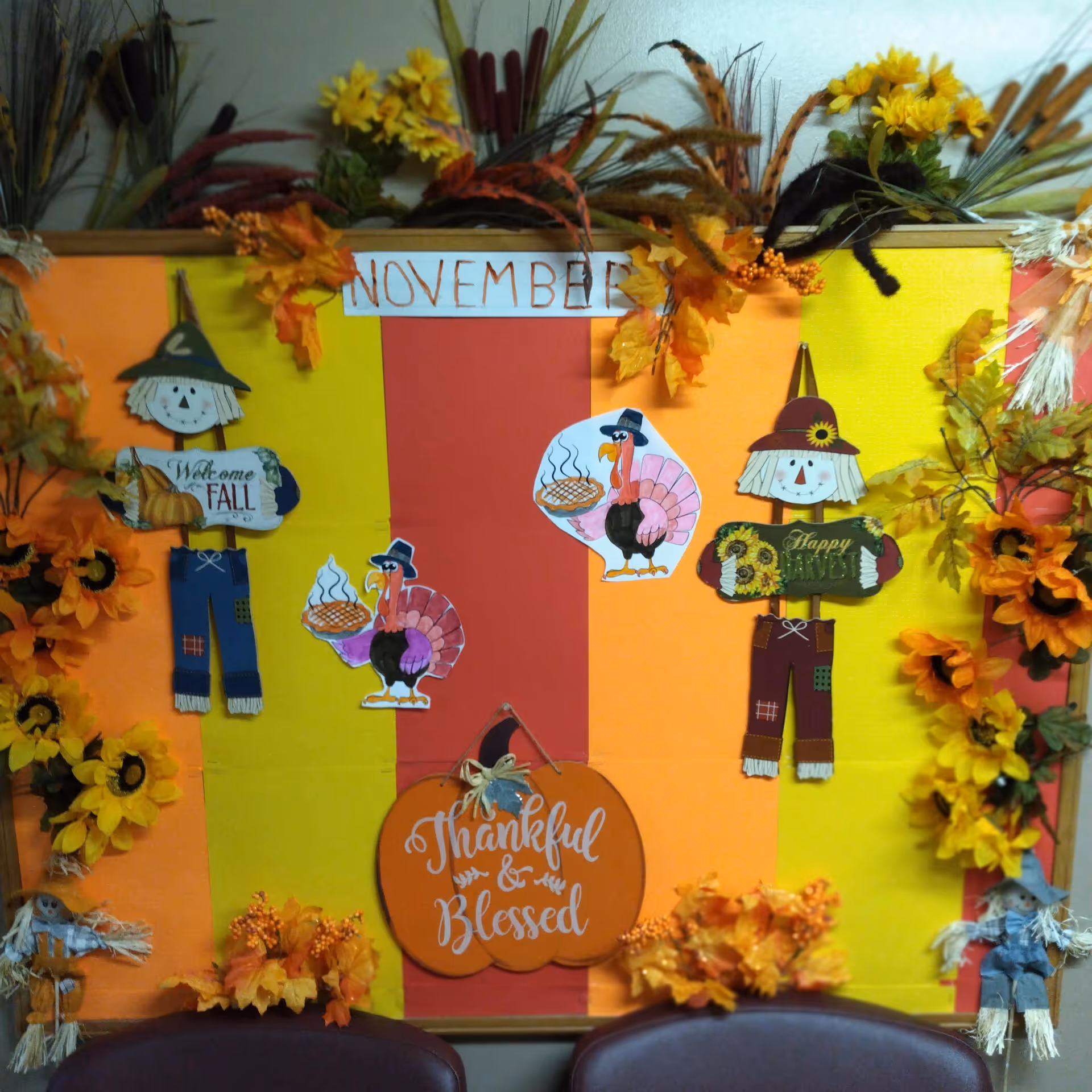 A colorful bulletin board decorated for November with fall-themed decorations including scarecrows holding signs that say 'Welcome Fall' and 'Happy Harvest', two cartoon turkeys holding pies, and a pumpkin-shaped sign that reads 'Thankful & Blessed'. The board is bordered with artificial autumn leaves and flowers, and there are two chairs partially visible at the bottom.