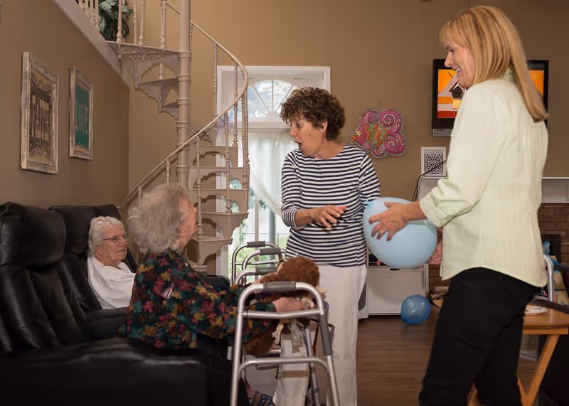 A group of elderly women and two caregivers are interacting in a cozy living room with a spiral staircase in the background. One caregiver is holding a blue balloon while the other gestures animatedly. Two elderly women are seated, one with a walker and a stuffed animal on her lap.