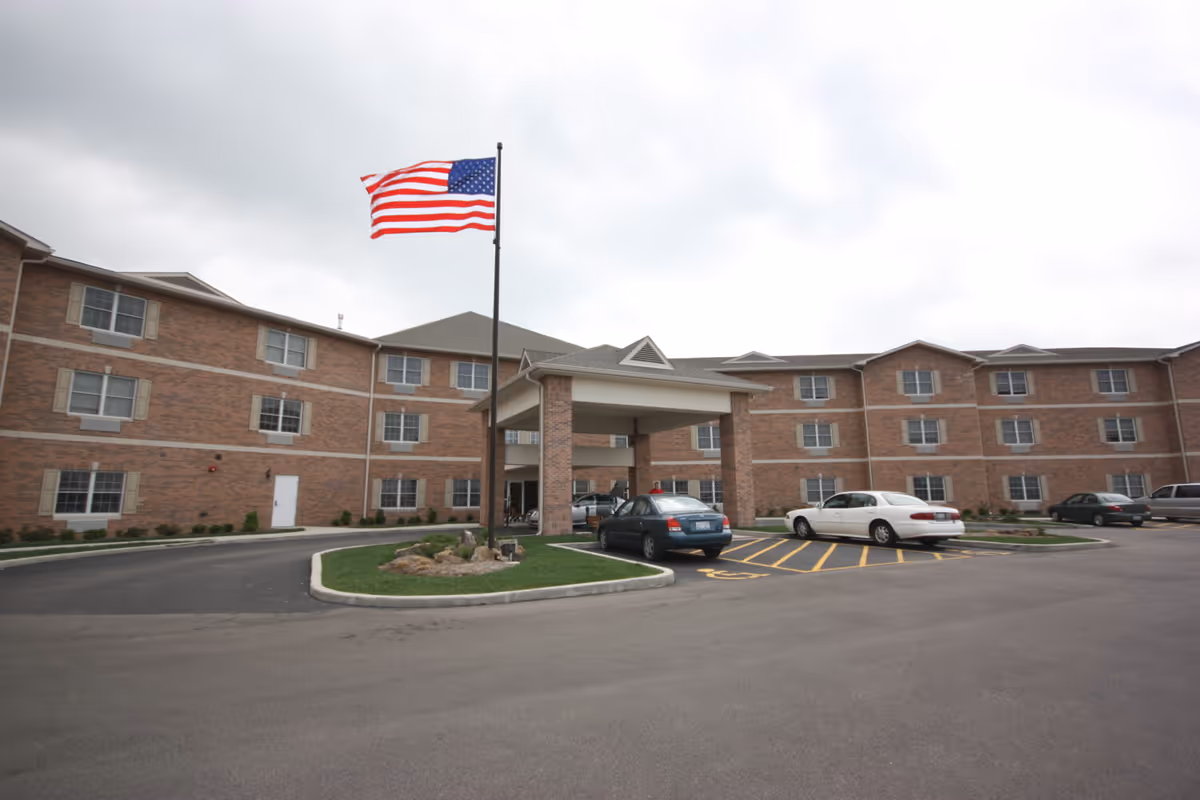 Exterior view of a three-story brick building with multiple windows, a covered entrance, and an American flag on a flagpole in front. Several cars are parked in the parking lot, including a handicapped parking space near the entrance.
