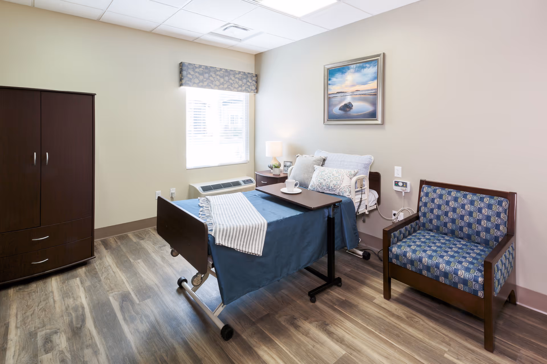 A clean and well-lit senior living bedroom with a hospital-style bed covered with a blue blanket and a striped throw. There is a small table over the bed with a cup and saucer. Next to the bed is a nightstand with a lamp and a small plant. A patterned armchair is placed against the wall, and a large wooden wardrobe stands near a window with blinds and a decorative valance. A framed landscape painting hangs on the wall above the bed.