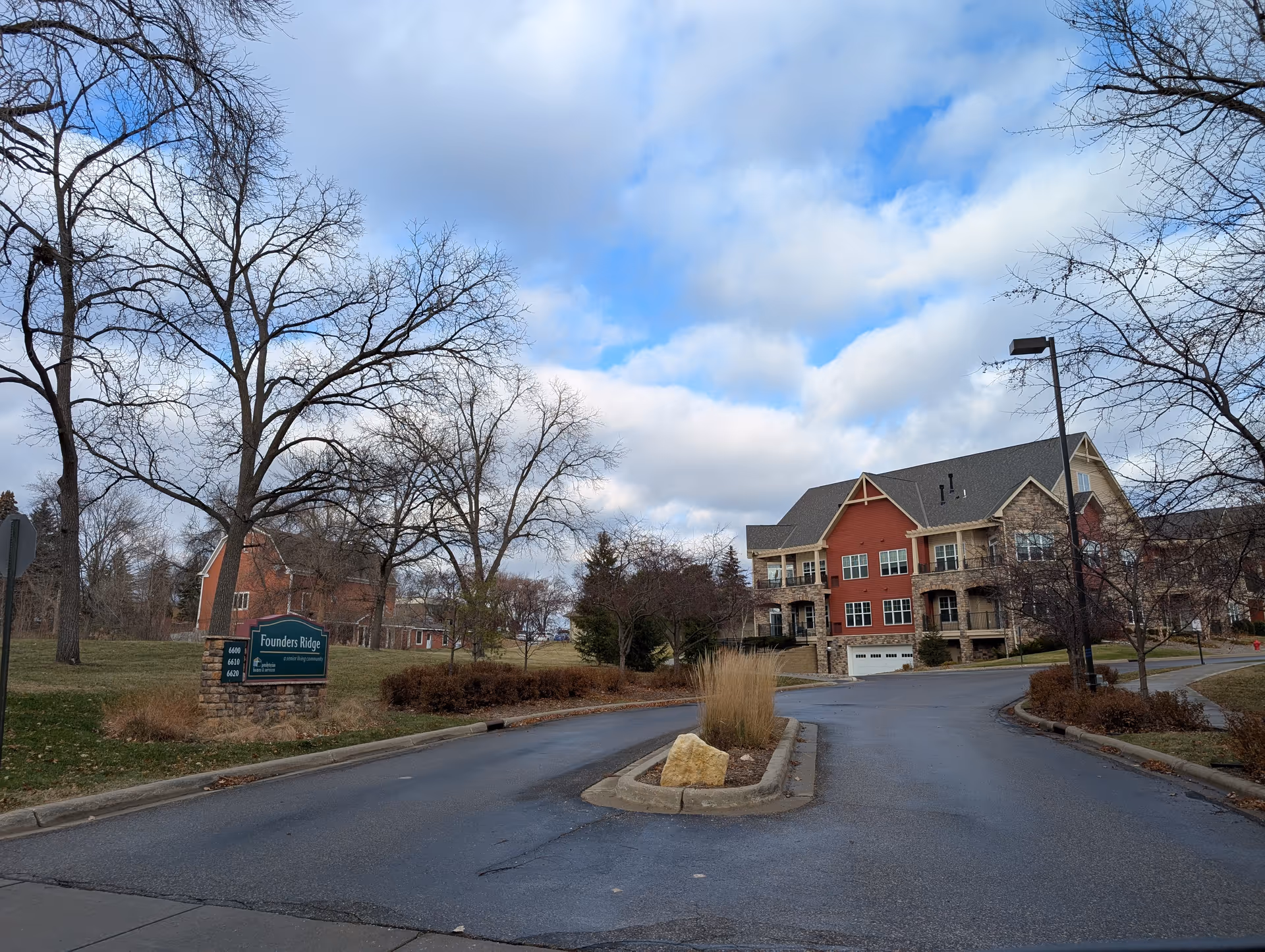 View of the entrance road to Founders Ridge senior living facility with a sign on the left side and a multi-story residential building in the background. Leafless trees and a partly cloudy sky are visible.