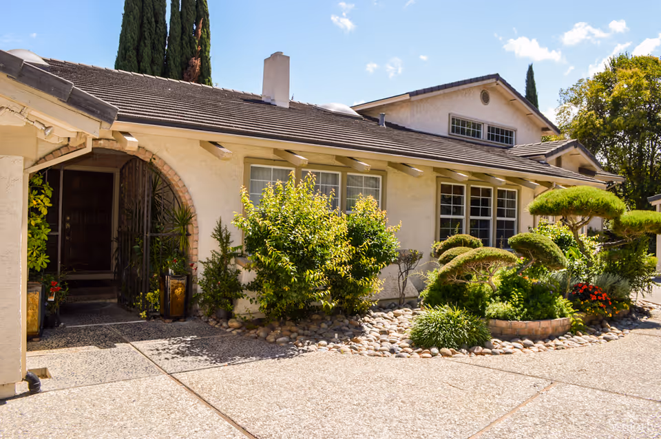 Front exterior of a single-story house with an arched entrance, landscaped shrubs, and a paved driveway.