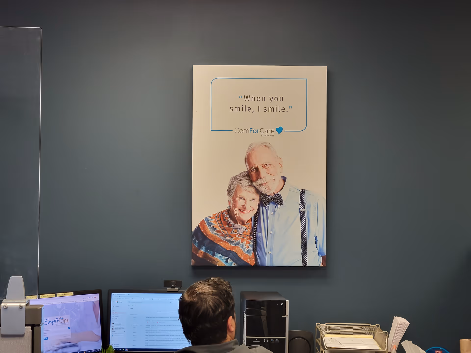 Office reception area with teal wall, large poster of smiling elderly couple, and computer desks.