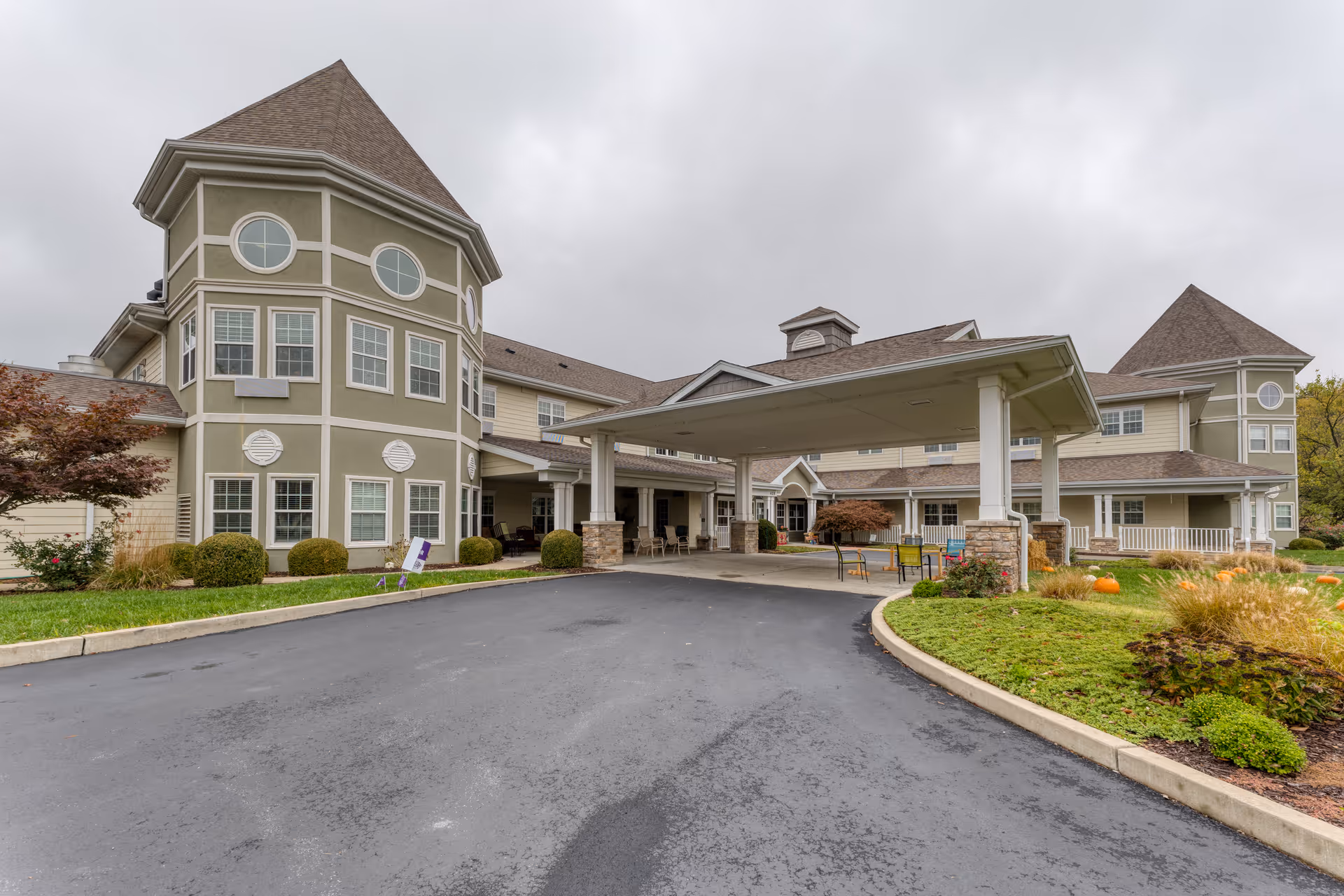 Exterior view of a senior living facility with a large covered entrance, multiple windows, and two prominent turret-like structures. The driveway is paved and curves around landscaped areas with bushes, small trees, and pumpkins. The sky is overcast.