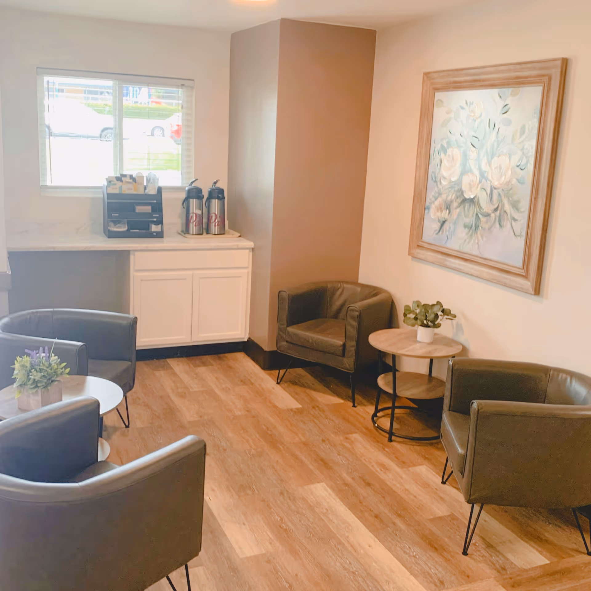 Interior waiting area with brown chairs, small tables, coffee station, and wall art.