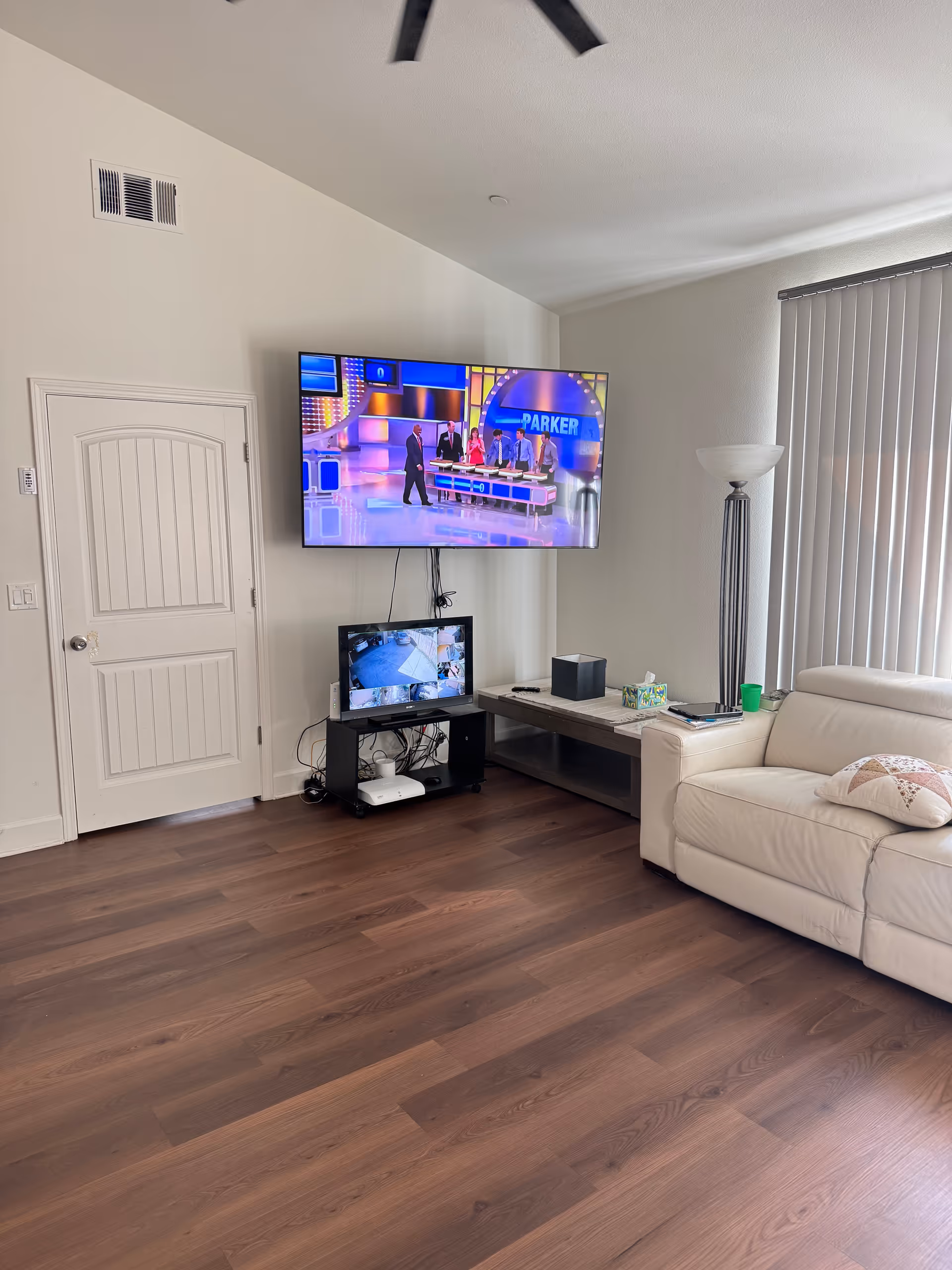 Living room with mounted TV, smaller monitor, sofa, floor lamp, and blinds.
