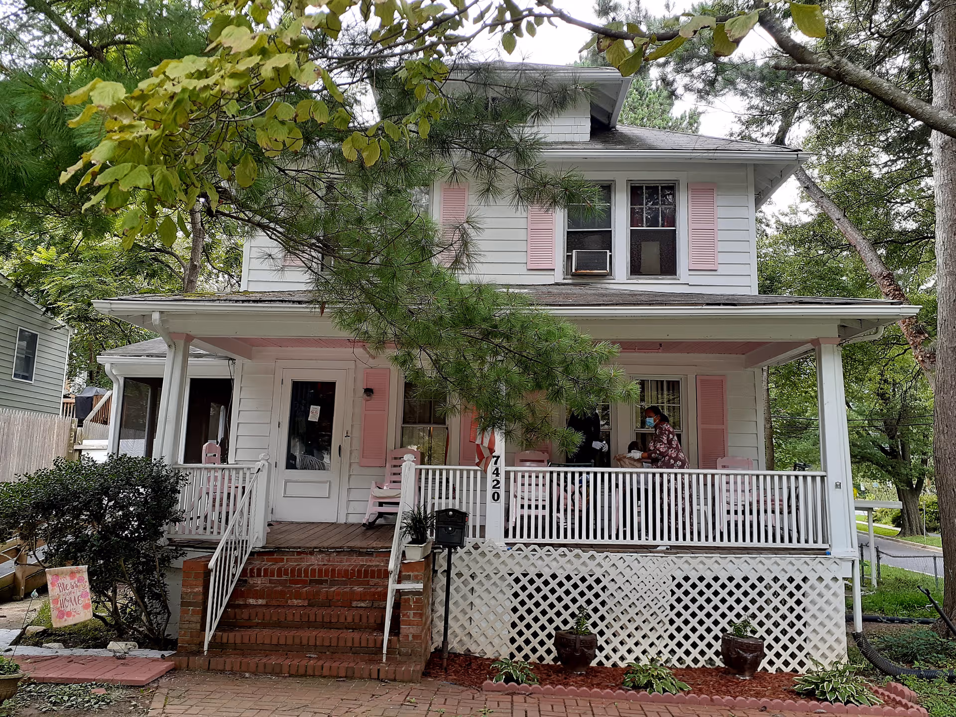 Two-story white house with pink shutters and a wraparound porch where residents sit.
