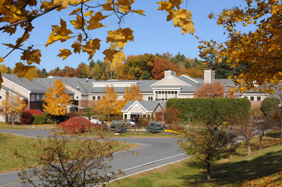 Exterior view of Kendal At Hanover senior living facility surrounded by trees with autumn foliage. The building has a light-colored facade with a gray roof and is set against a backdrop of more trees under a clear blue sky.