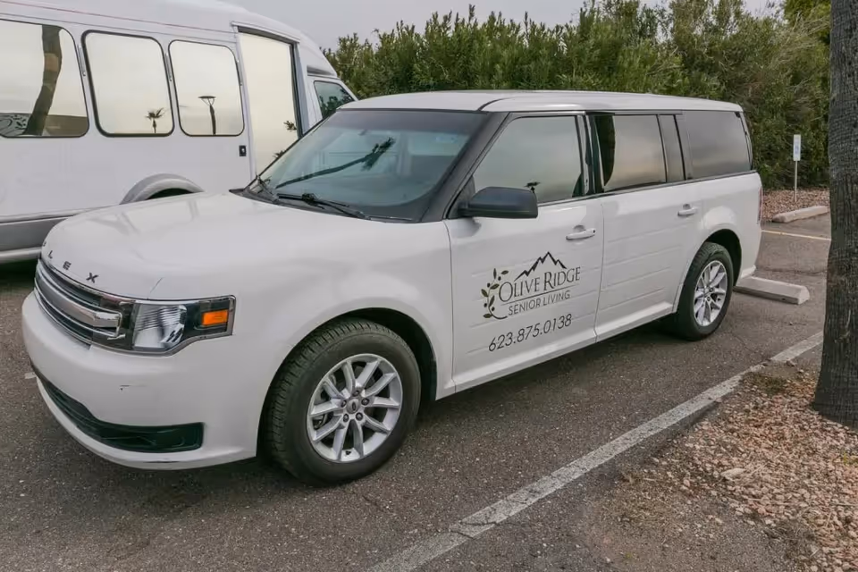 White passenger vehicle parked in a parking lot with the logo and contact number of Olive Ridge Senior Living on the side door. Another white vehicle is partially visible in the background.
