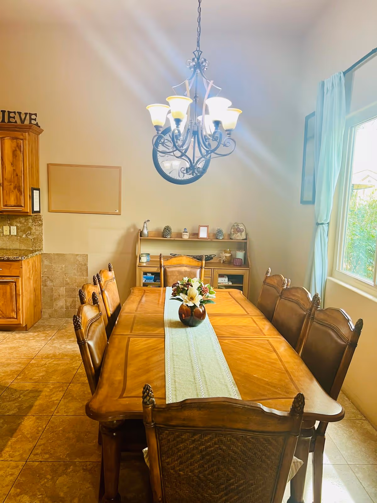 Wooden dining table with eight chairs, chandelier, and sideboard in a sunny dining room.