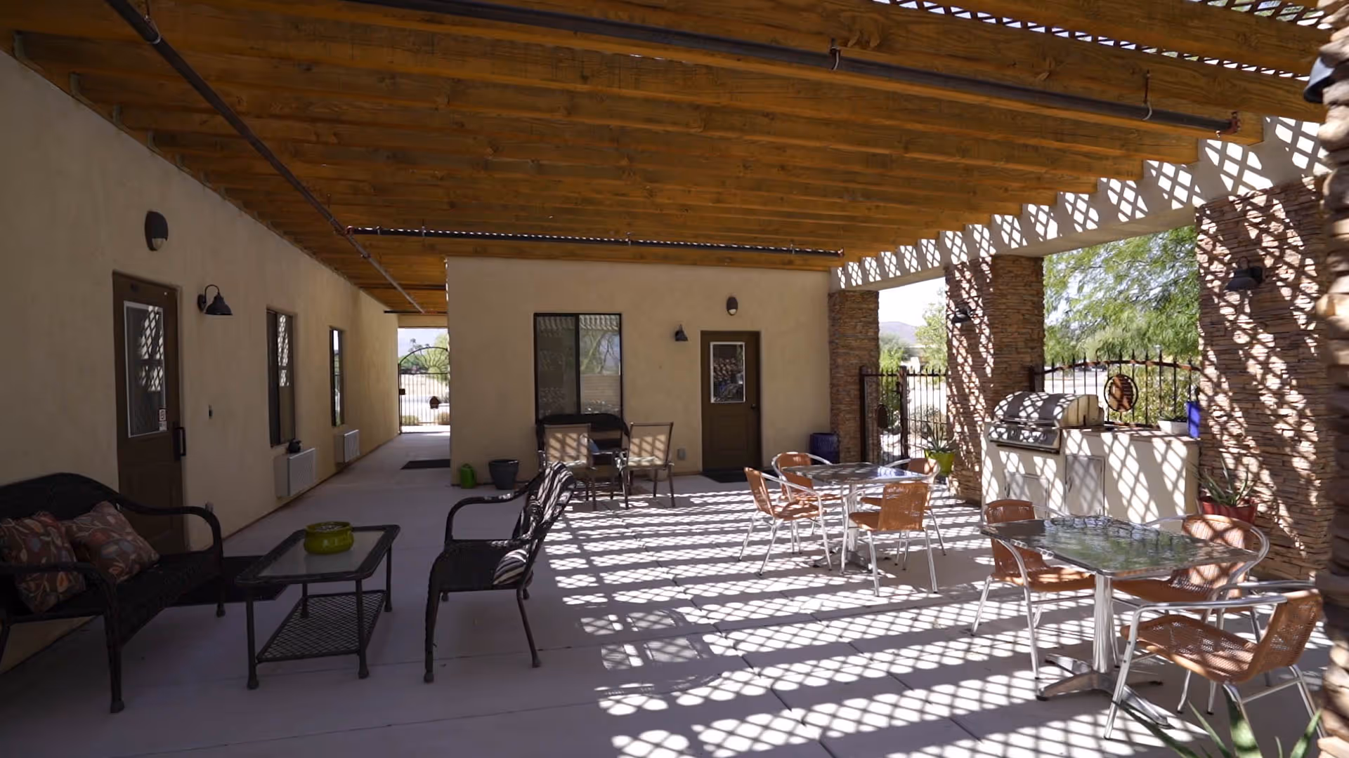 Shaded outdoor patio with tables, chairs, and a barbecue area beside beige building walls.