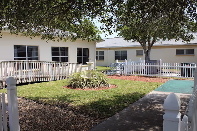 Outdoor courtyard area at Lexington Health and Rehabilitation Center featuring a white multi-tiered fountain surrounded by greenery, a white picket fence, benches, a tree providing shade, and a building with windows in the background.