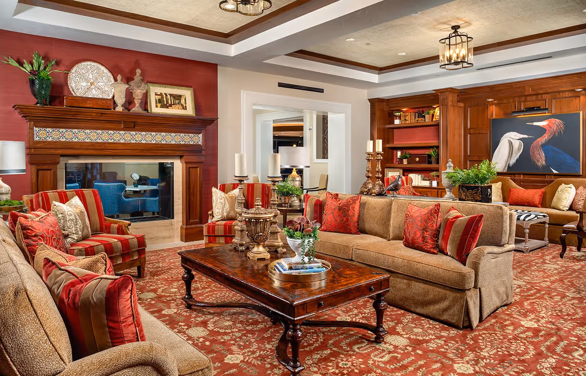 Well-appointed living room with sofas and striped armchairs arranged around a wooden coffee table on a patterned red rug, a fireplace and built-in shelving with artwork.