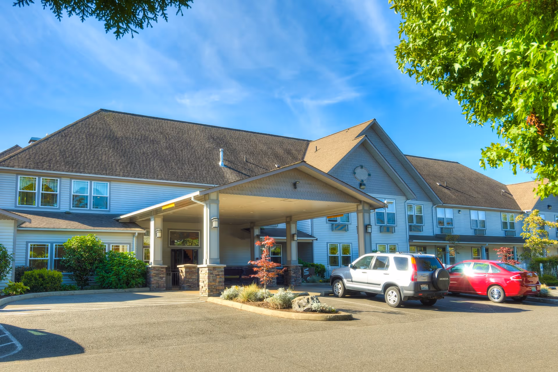 Front entrance of a senior living facility with a covered drop-off, landscaping, and parked cars.
