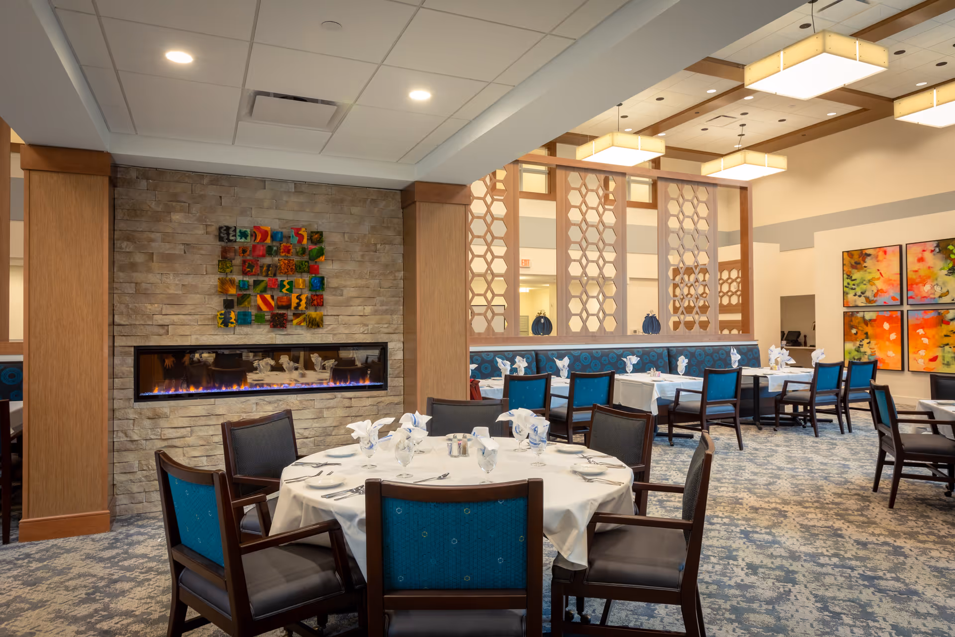 A well-lit dining room with round and rectangular tables set with white tablecloths, napkins, and silverware. The room features a modern fireplace set in a stone wall, decorative wooden partitions with a honeycomb pattern, blue cushioned chairs, and colorful abstract artwork on the walls.