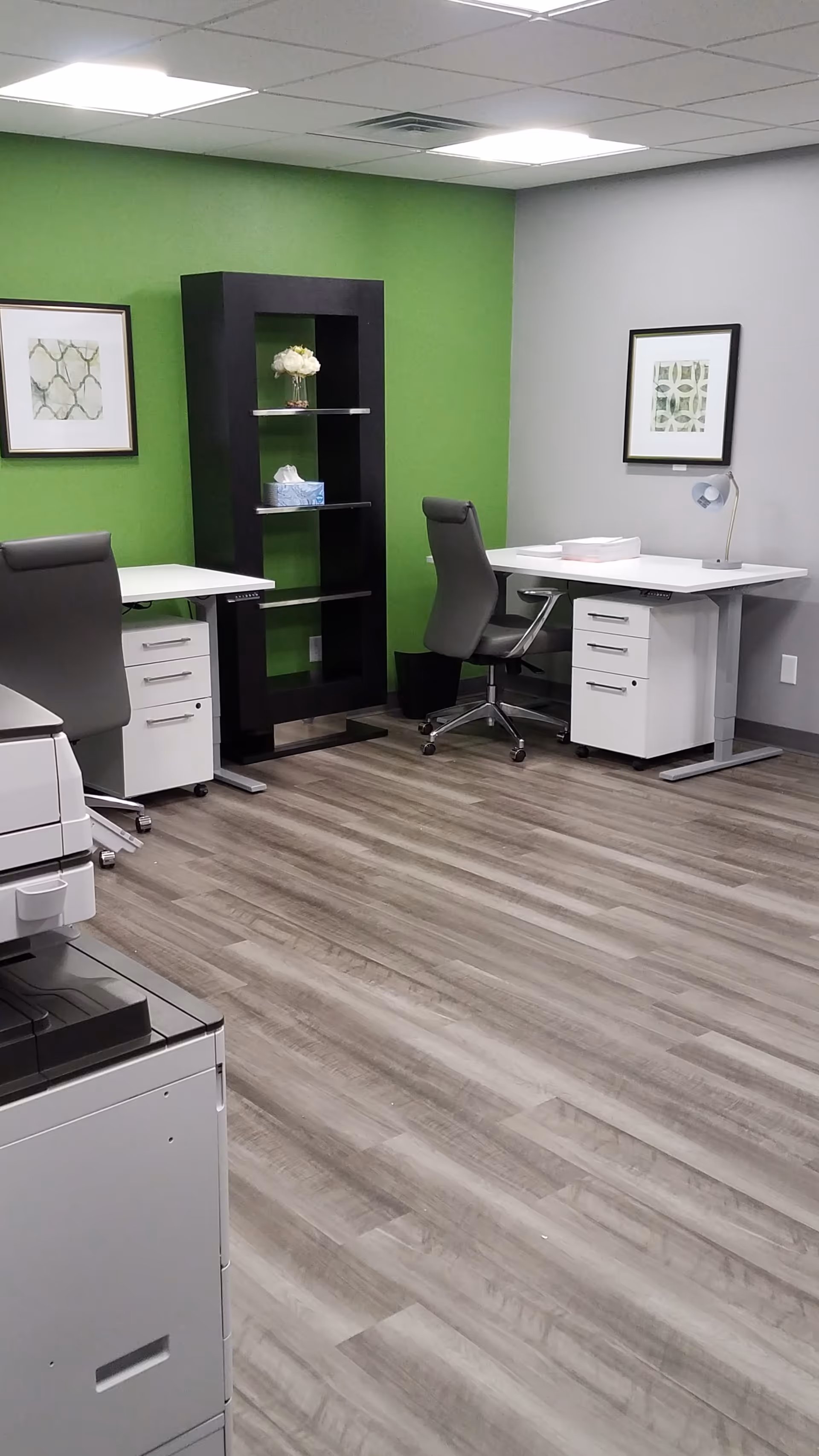 Office workspace with two desks, grey chairs, a black shelf, and green accent wall.