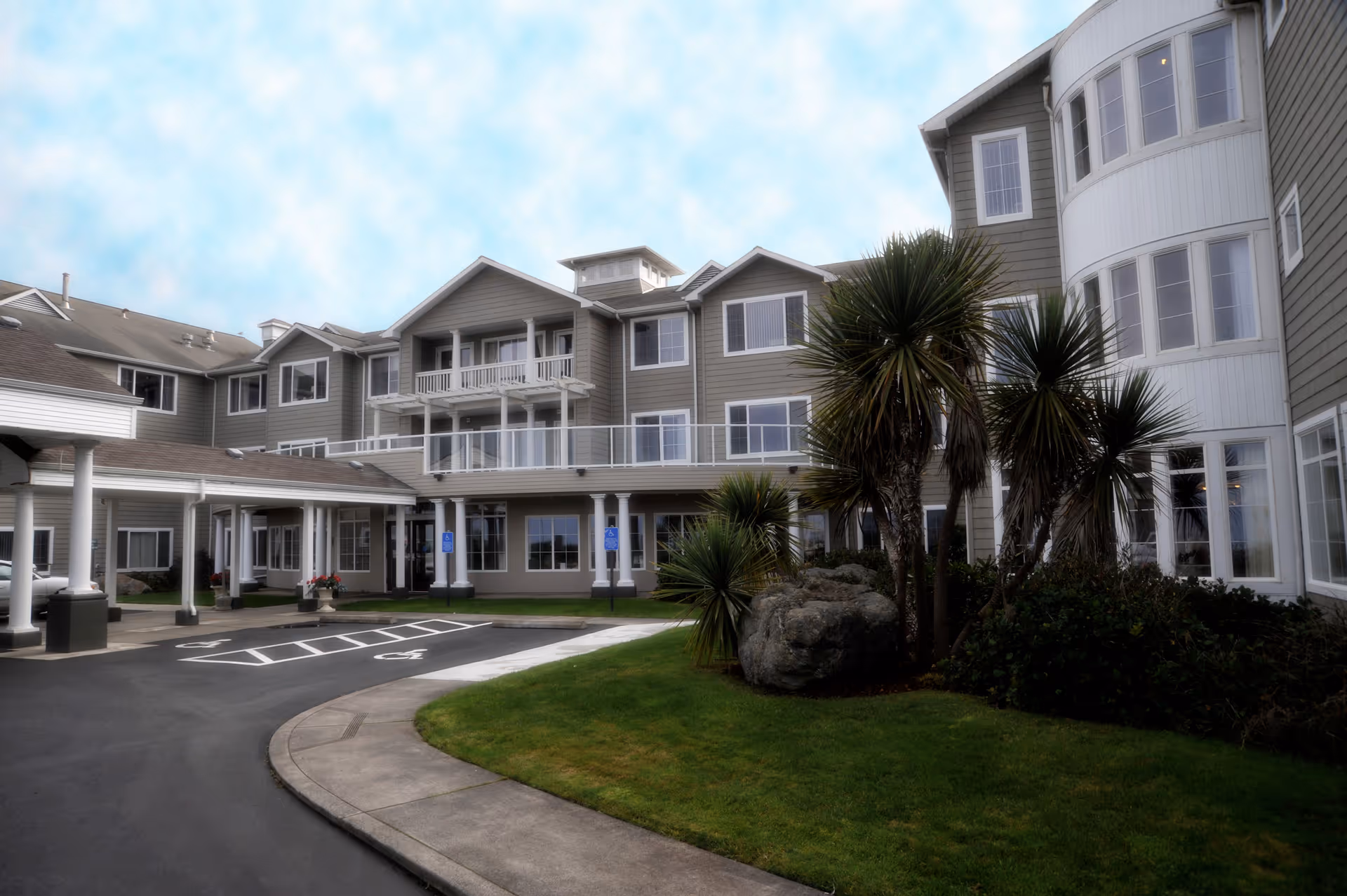 Exterior view of a multi-story senior living facility building with gray siding, white trim, and multiple windows. There is a covered entrance with columns, a driveway with handicap parking spaces, green lawn, and several palm-like trees and shrubs in the landscaped area.