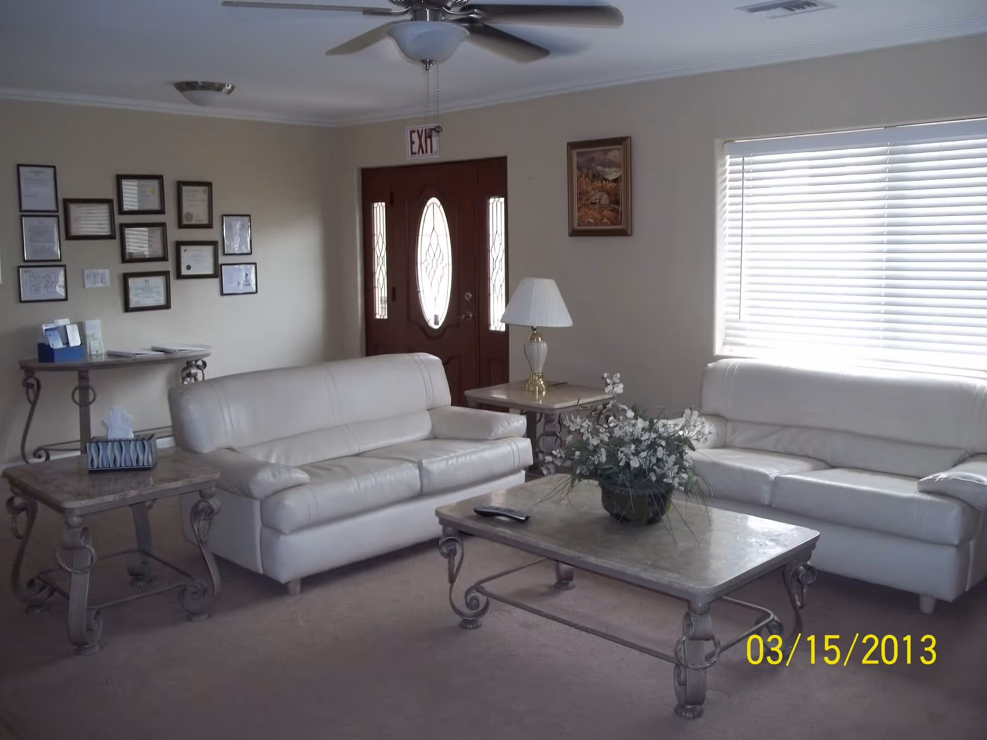 A living room with two white leather sofas facing each other, a coffee table with a floral arrangement in the center, two side tables with a lamp and tissue box, a ceiling fan, framed certificates on one wall, a window with blinds, and a wooden front door with glass panels.