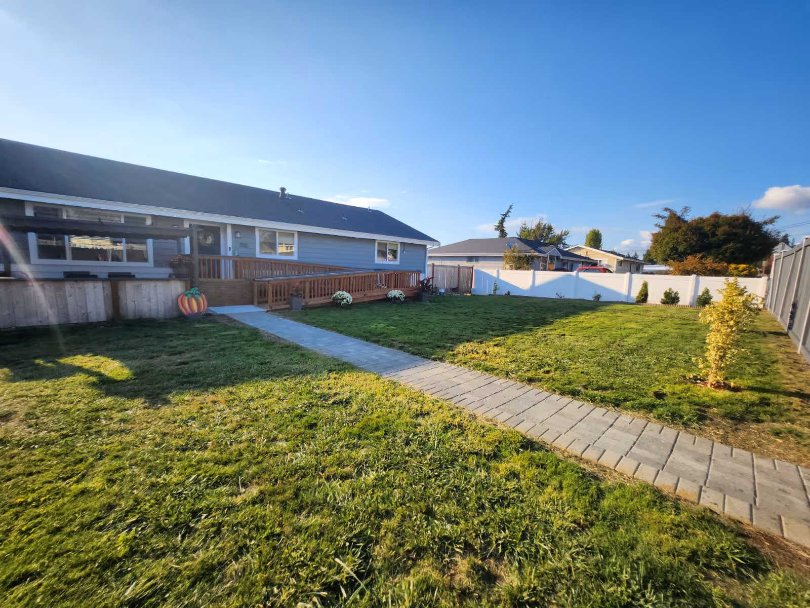 A large, well-maintained grassy backyard with a paved walkway leading to a single-story house with a wooden ramp. The yard is enclosed by a wooden fence on the left and a white vinyl fence at the back. There are a few small plants and shrubs along the fences, and the sky is clear and blue.