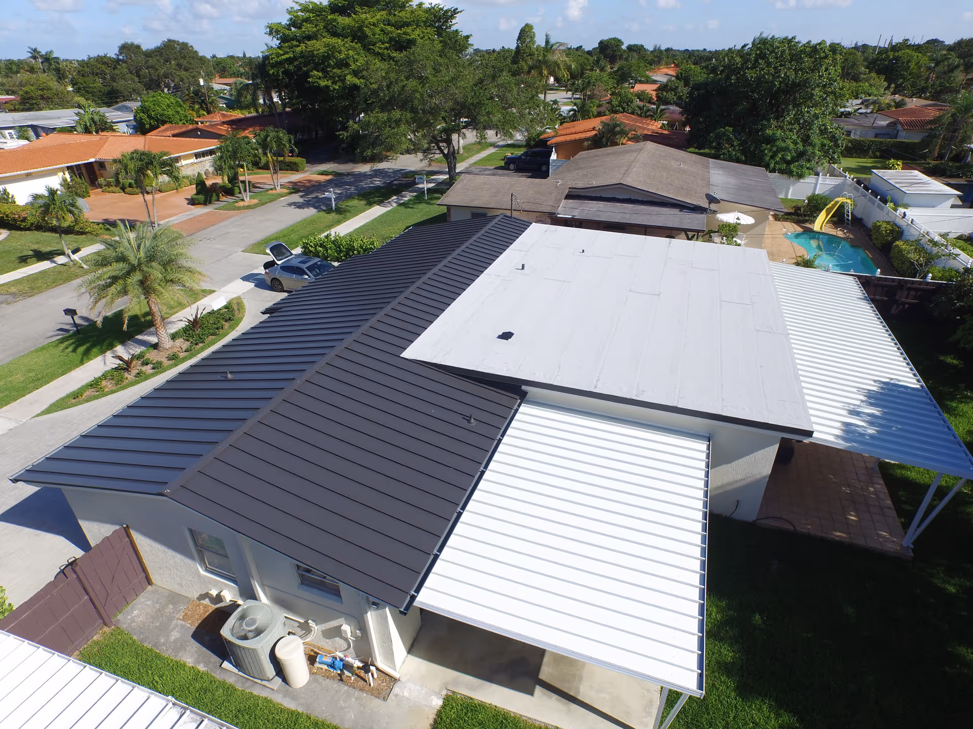 Aerial view of a residential neighborhood showing the rooftops of several houses, including a house with a dark metal roof and white awnings. The area has green lawns, palm trees, and a swimming pool with a yellow slide in one backyard.