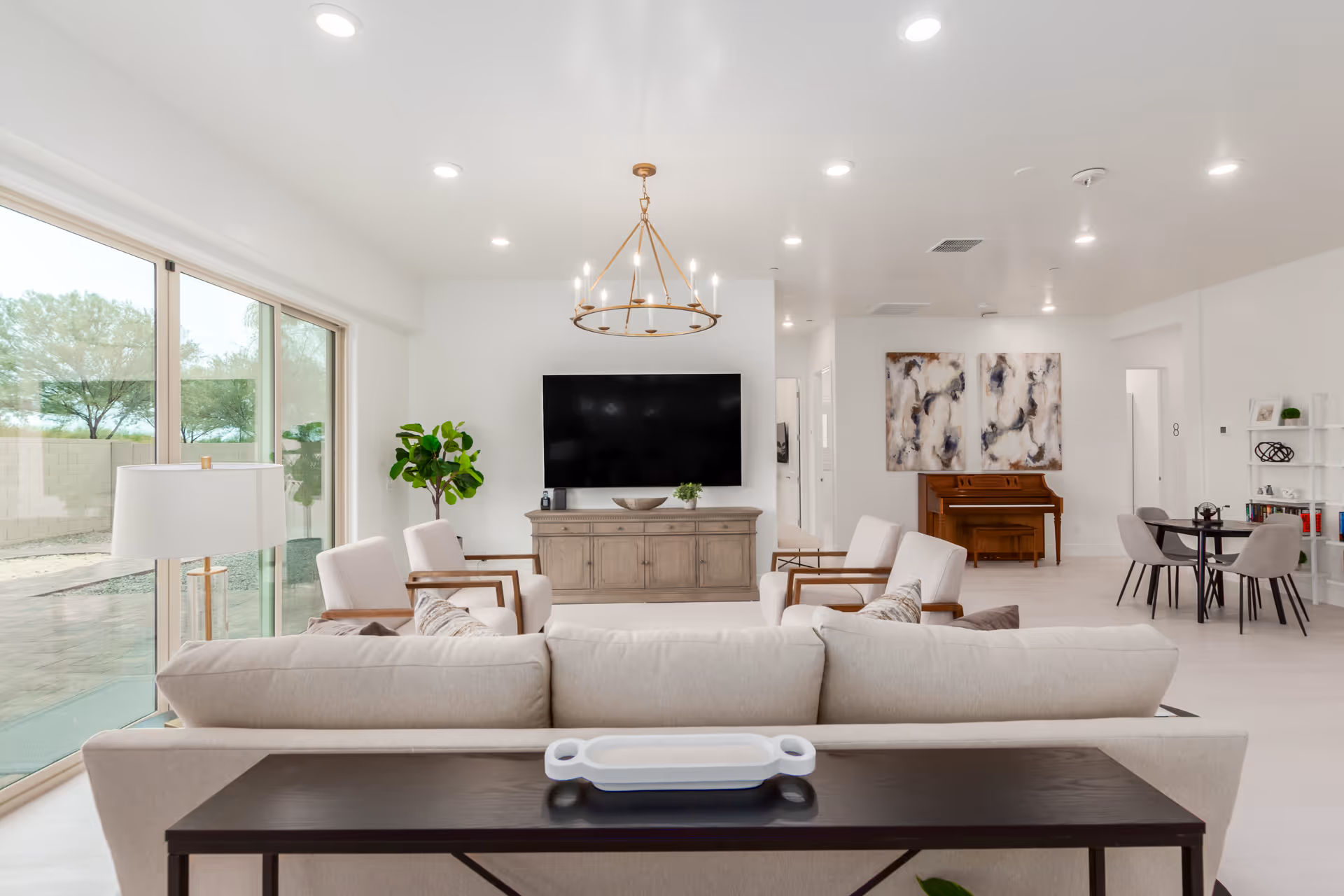 Bright, modern living room with seating area, TV, piano, and dining nook in a senior facility.