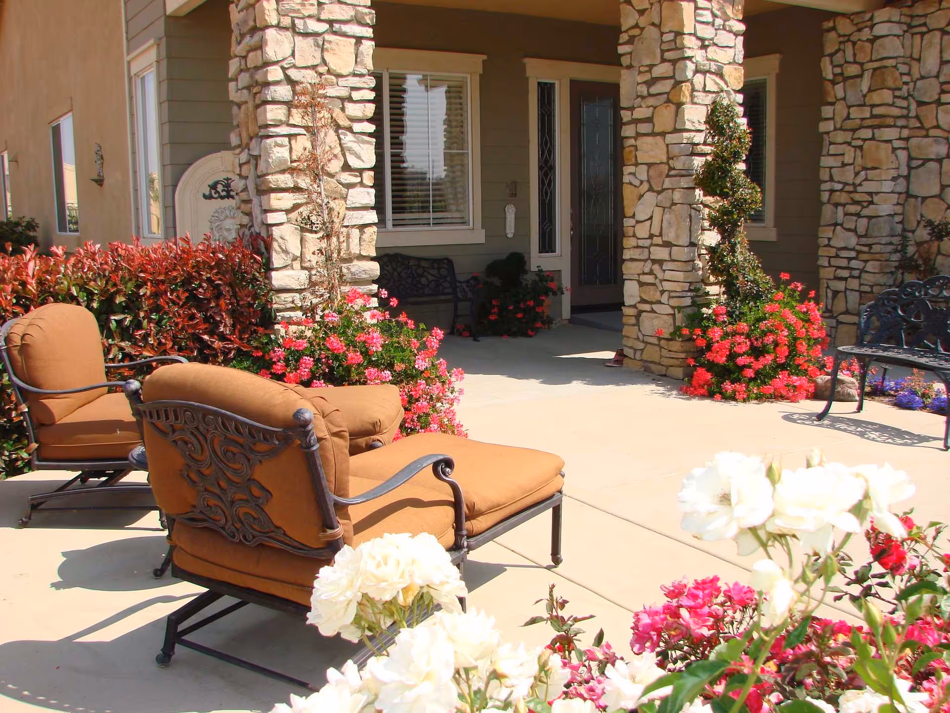 Stone pillars frame a porch with brown cushioned chairs and blooming flowers outside.