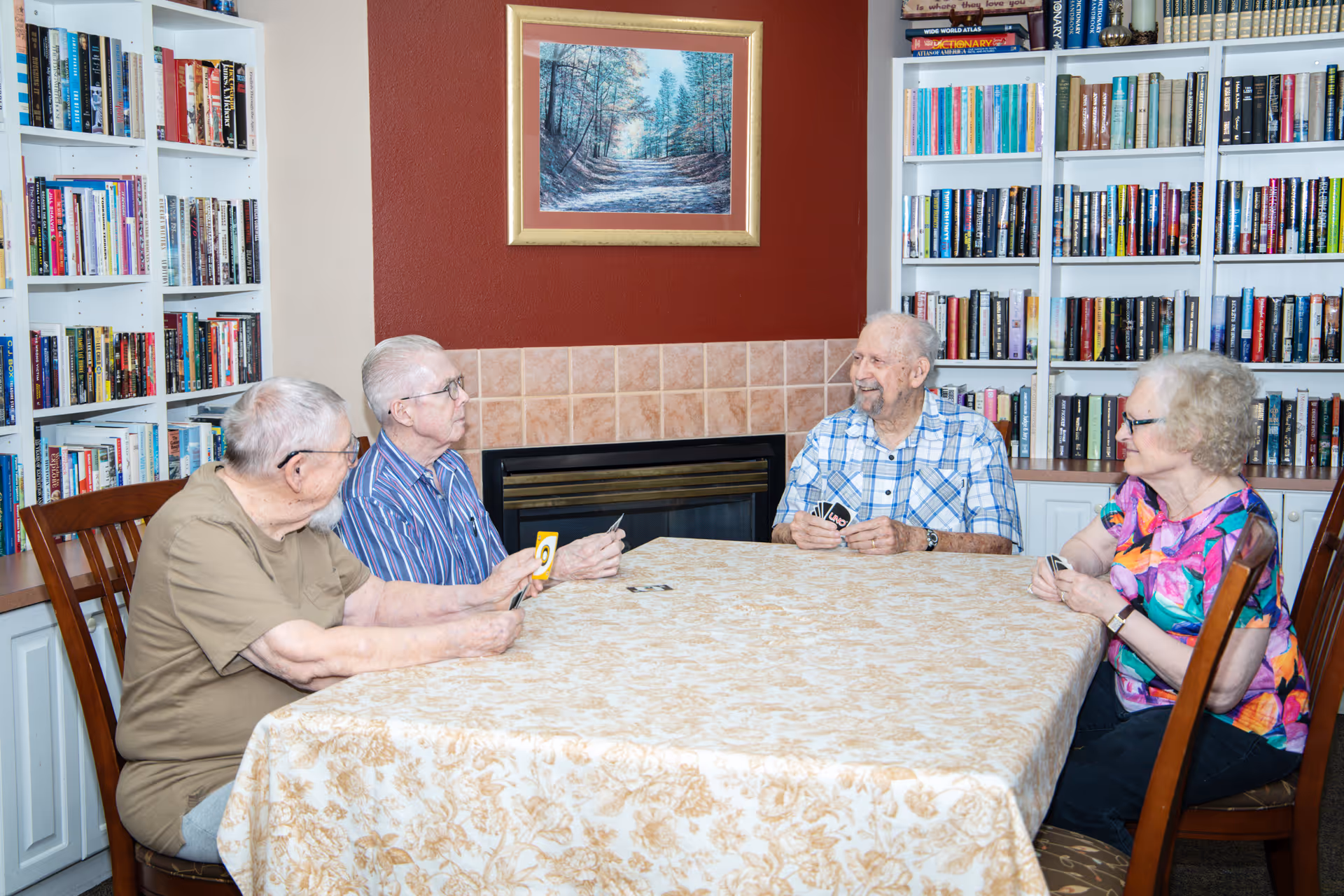 Four elderly people sitting around a table playing cards in a room with bookshelves filled with books and a fireplace with a framed painting above it.