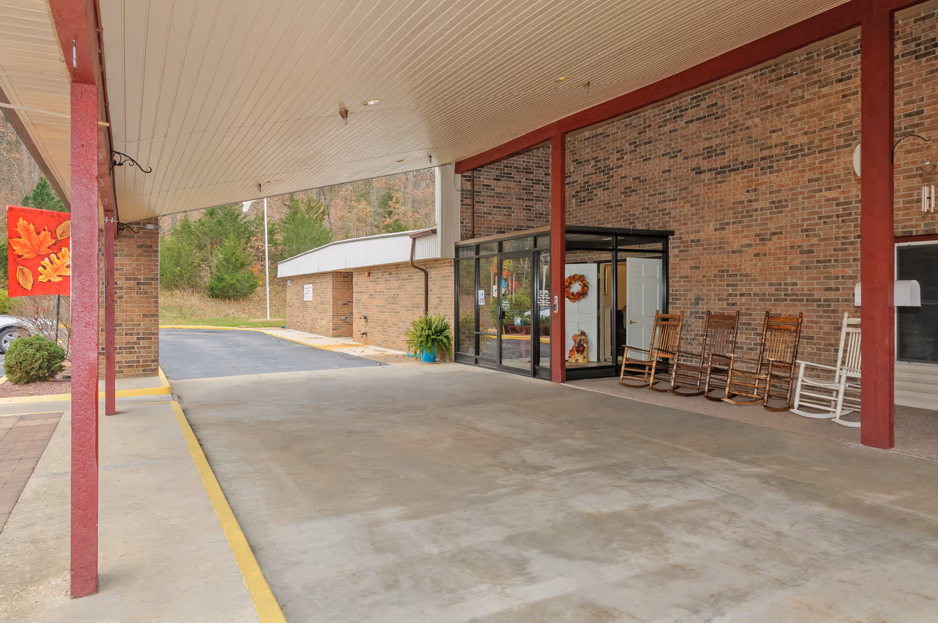 Covered entrance area of a brick building with a glass door entrance. Several wooden rocking chairs are lined up against the wall near the entrance. There is a red flag with autumn leaves hanging from a post on the left side. Trees and a paved driveway are visible in the background.