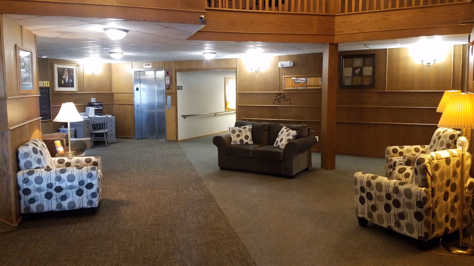 A cozy interior common area with wood-paneled walls, carpeted floors, and ceiling lights. The space features a brown loveseat with two patterned pillows, two patterned armchairs with a floor lamp between them, and a small desk with a chair near an elevator. There are framed pictures on the walls and a mirror above the loveseat.