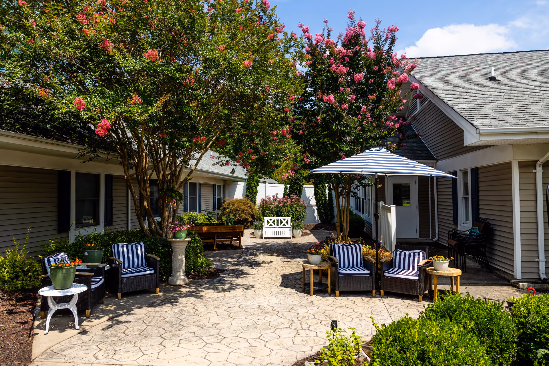Outdoor courtyard area at Churchland Nursing Home with paved stone walkway, several seating arrangements including wicker chairs with blue and white striped cushions, a striped umbrella, potted plants, flowering trees, and bushes under a partly cloudy blue sky.