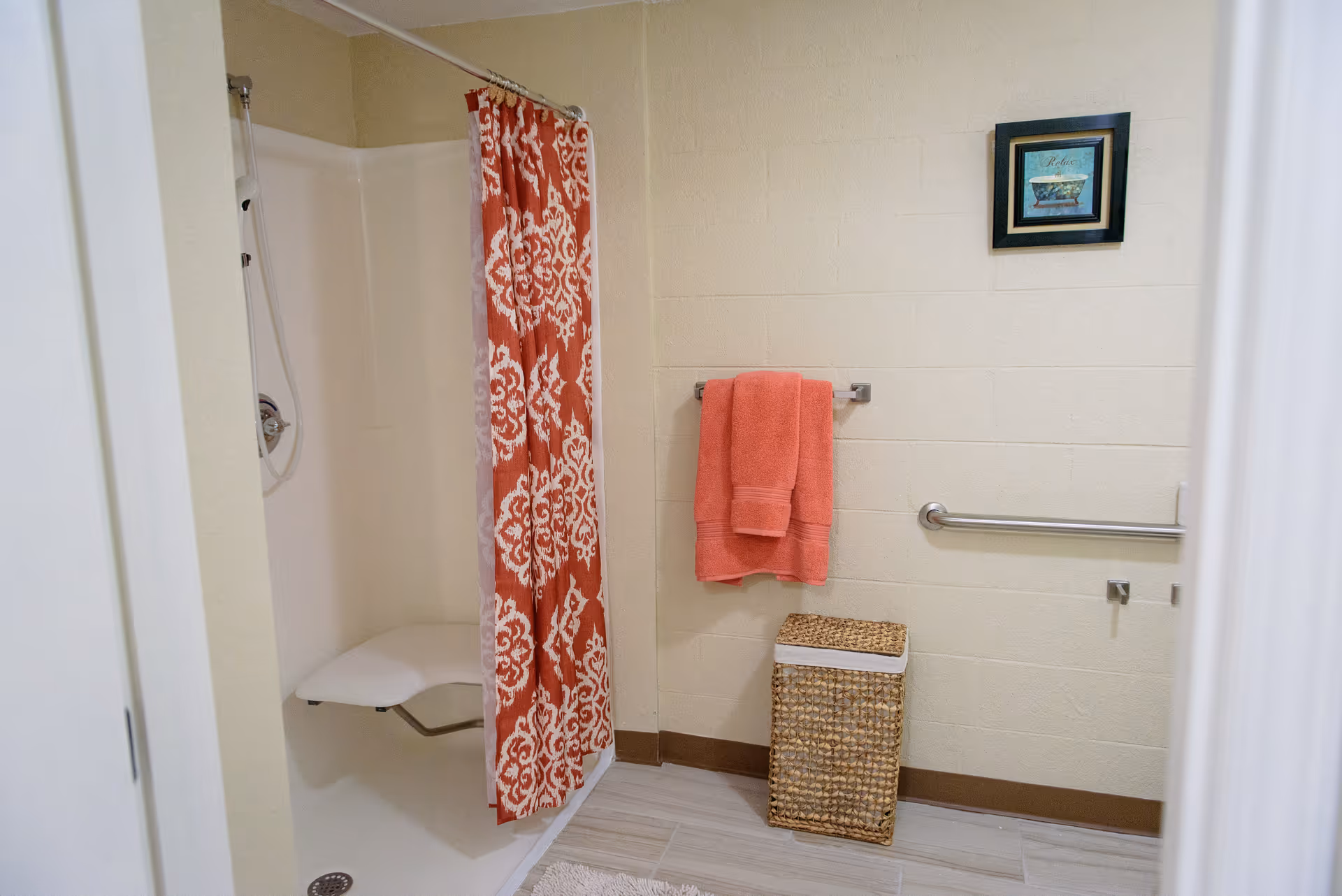 A bathroom with a walk-in shower featuring a white fold-down seat and a red and white patterned shower curtain. On the wall, there is a towel rack with two coral-colored towels, a grab bar, a wicker laundry basket, and a framed picture with a bathtub and the word 'Relax'.