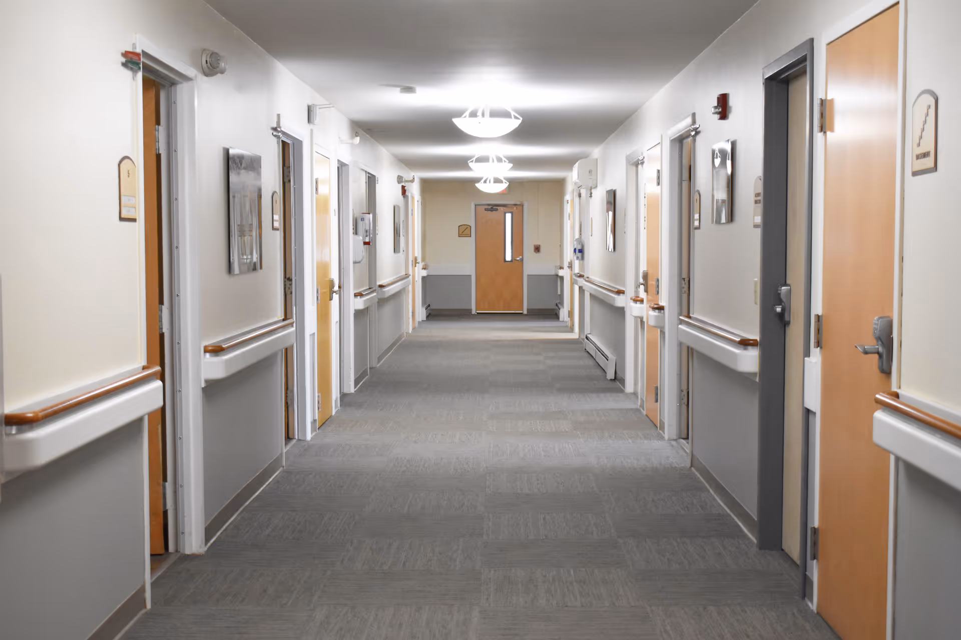 Long hallway in a senior living facility with doors, handrails, and bright ceiling lighting.