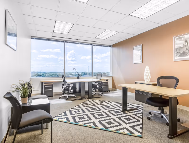 Bright office workspace with two desks, chairs, and a large window view.