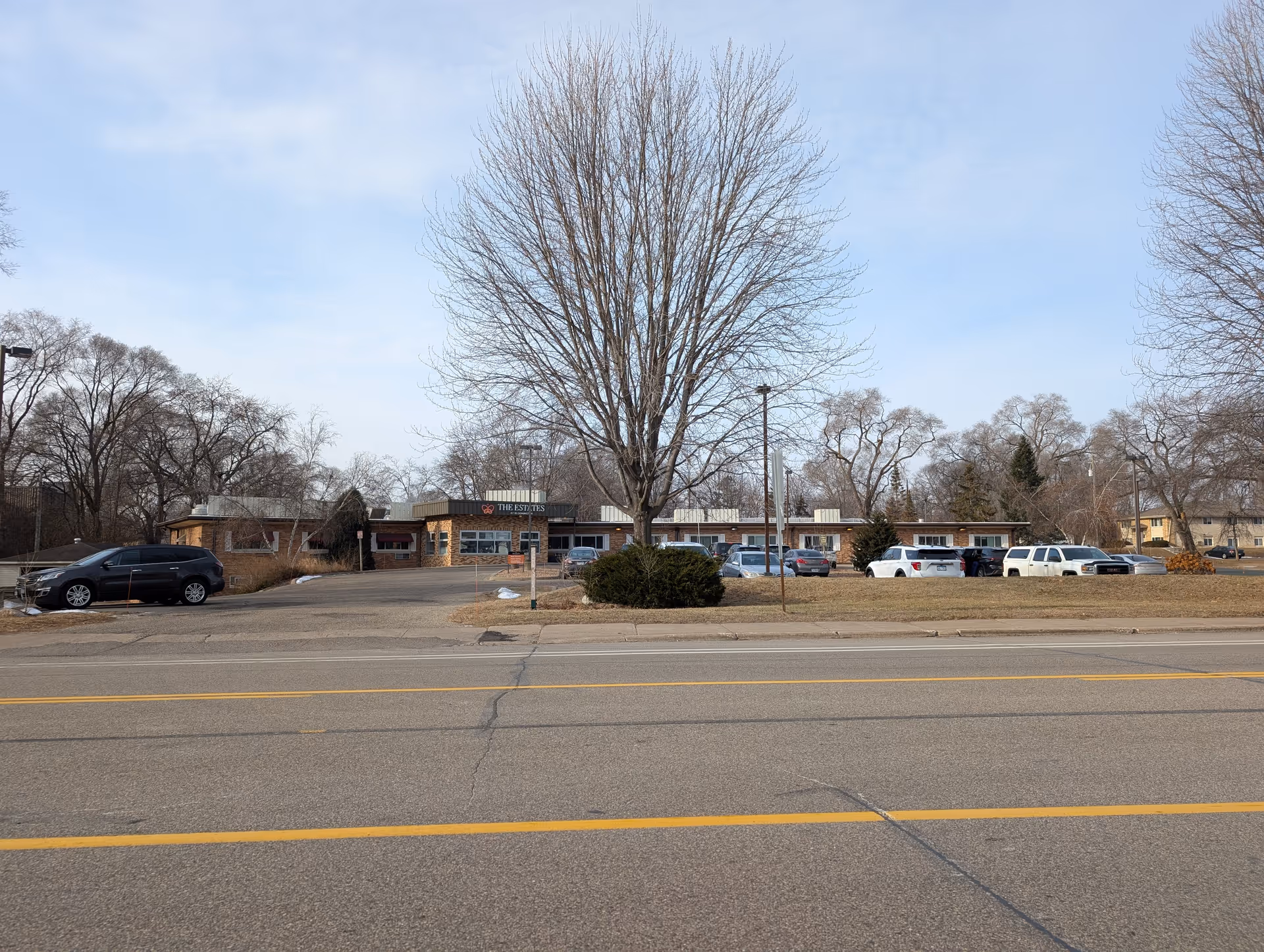 Front exterior of The Estates at Bloomington, a single-story brick building with a large leafless tree and parked cars in the driveway.