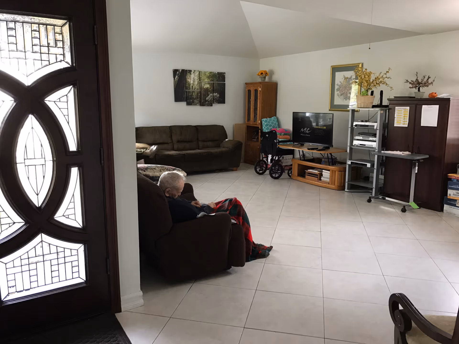 Living room with an elderly man seated in a chair, wheelchair nearby, TV and furniture.
