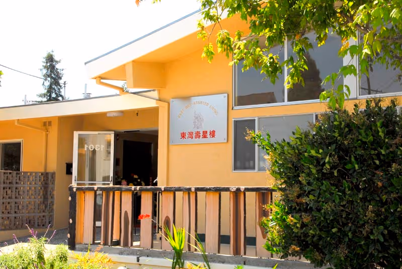 Entrance of East Bay Assisted Living showing a yellow building facade, main door, sign with English and Chinese text, and a small fenced porch with shrubs.