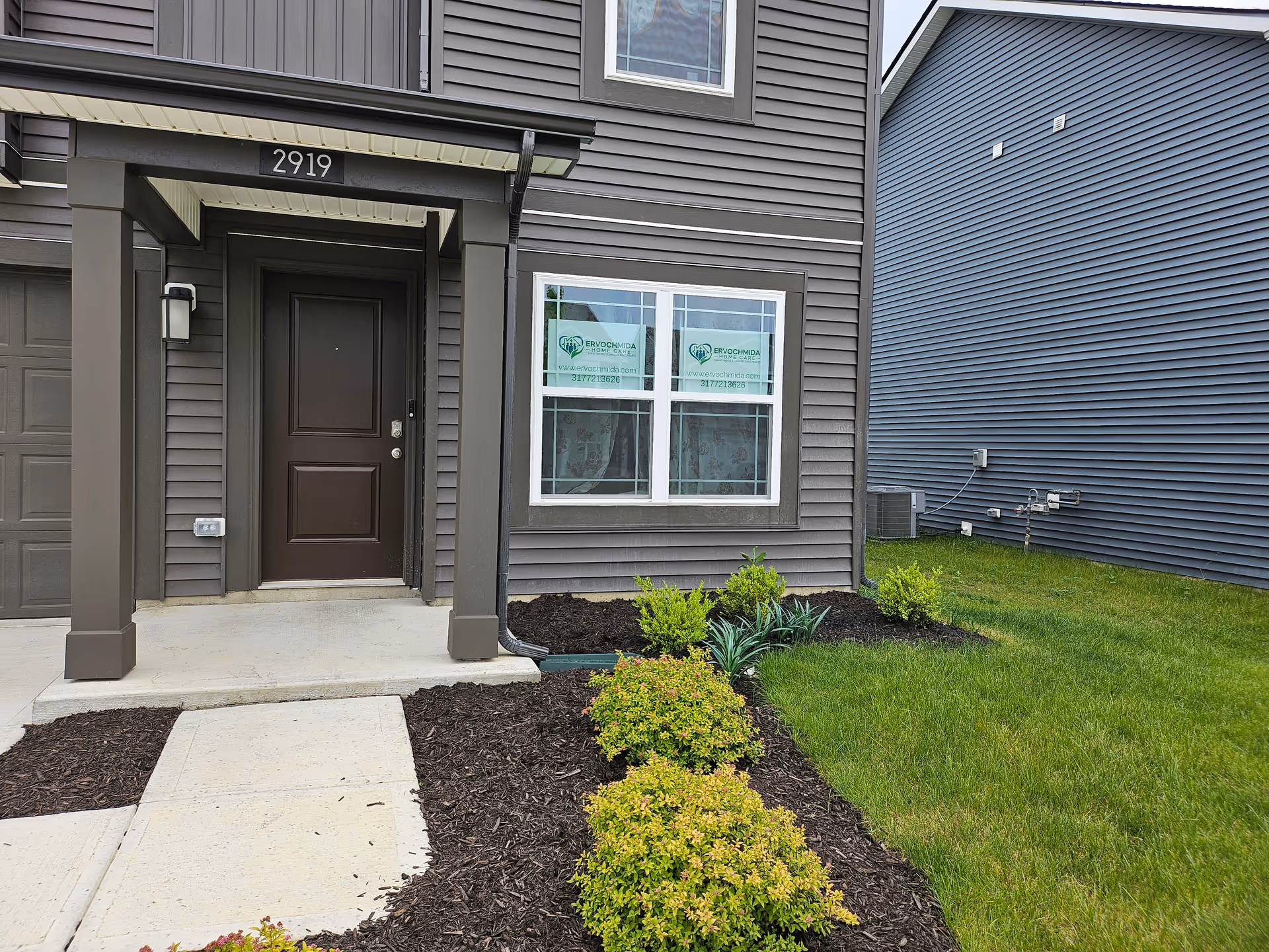 Front entrance of a brown-siding senior home with walkway and landscaped garden.