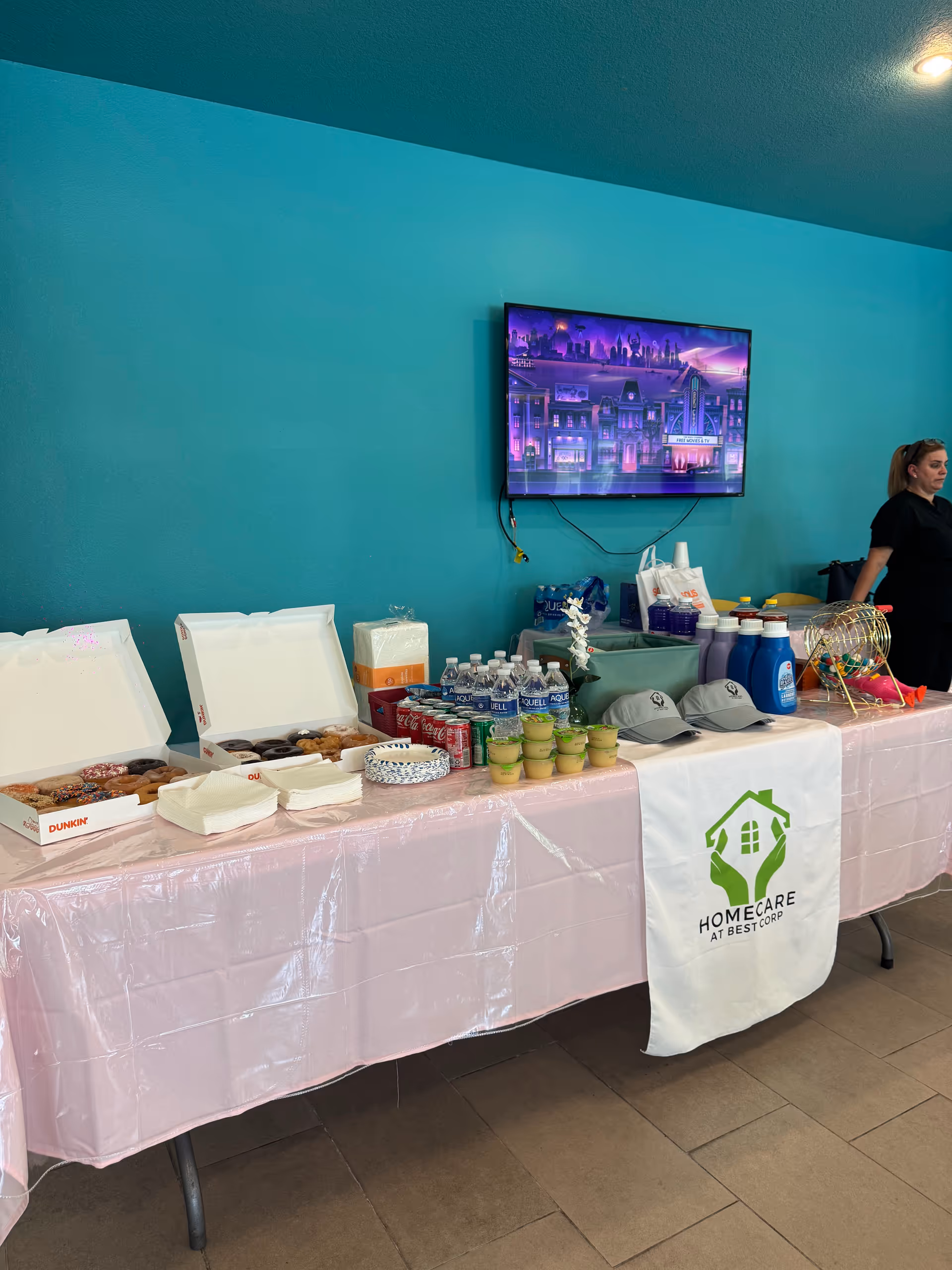 A long buffet table with pastries and drinks under teal walls and a TV.