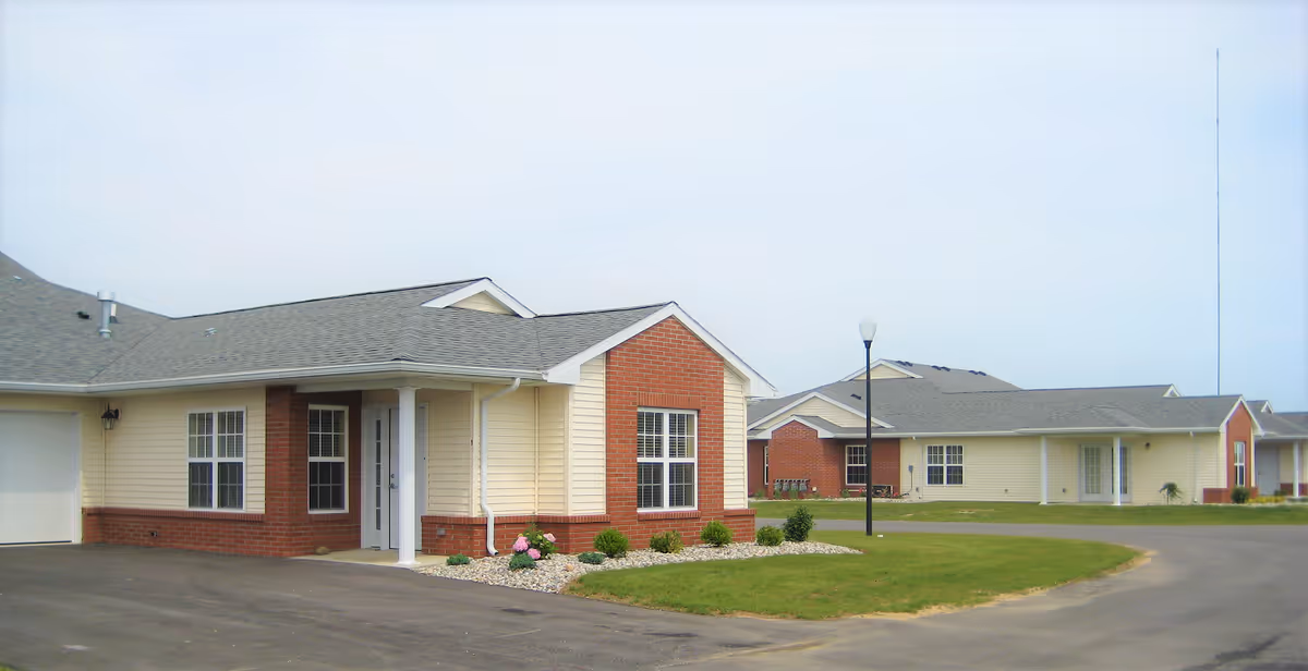 Exterior view of single-story residential buildings with beige siding and red brick accents, surrounded by a paved driveway and green lawn under a clear sky.