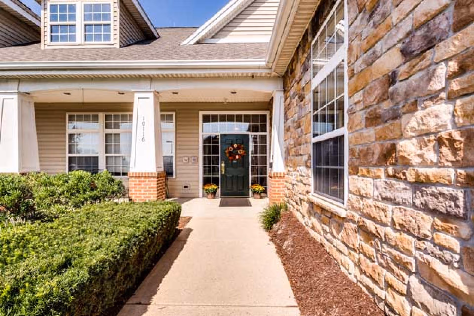 Front entrance of a building with a green door decorated with a fall wreath, flanked by two potted plants with yellow flowers. The building features beige siding, brick pillars, and a stone wall on the right side. There are trimmed bushes along the walkway leading to the door.