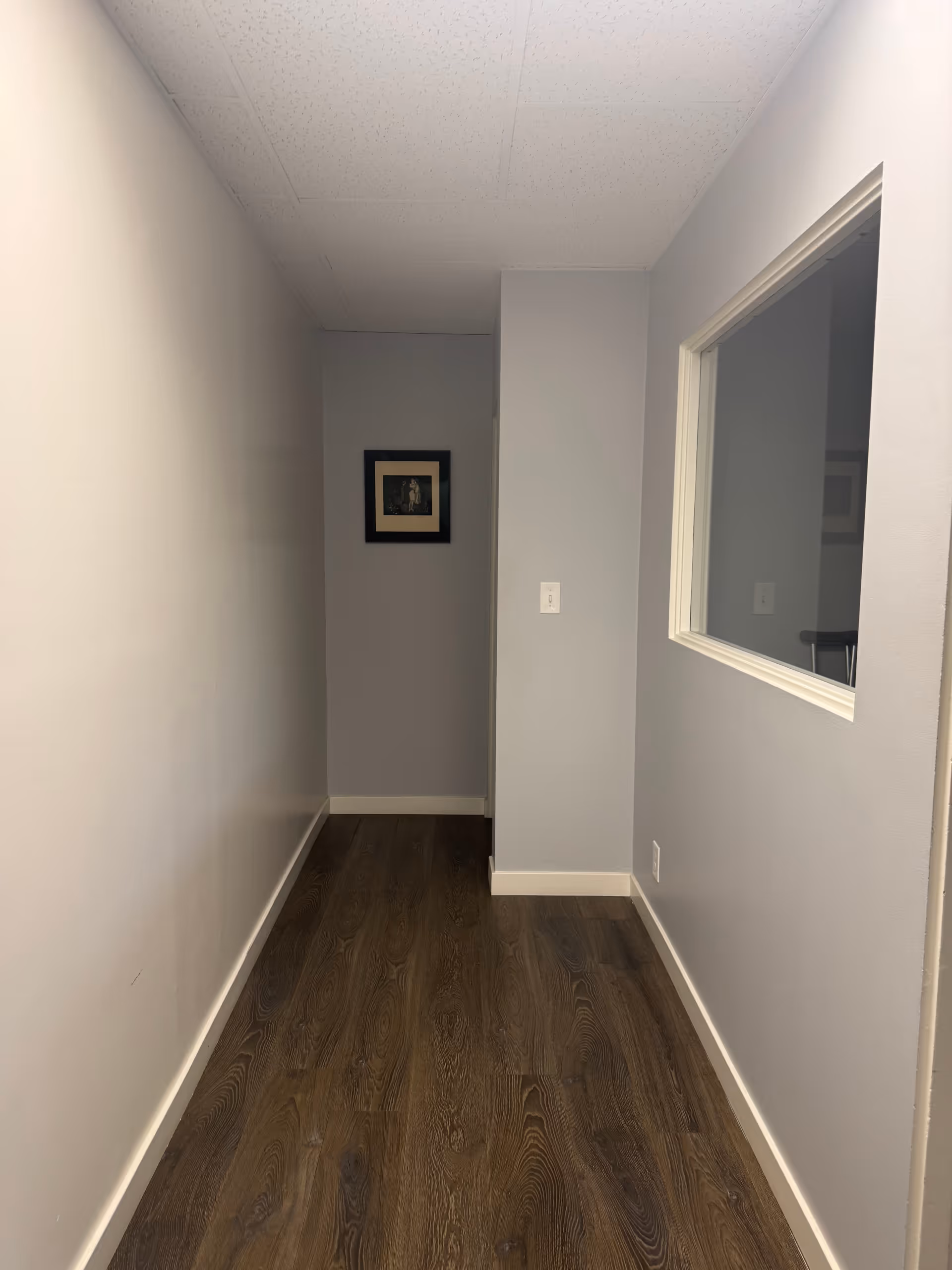 Narrow hallway with gray walls, wood flooring, framed art, and a window opening to a room.