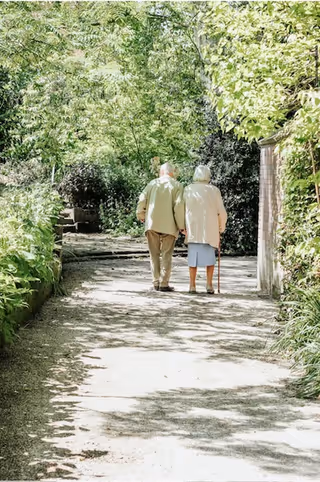 Two elderly individuals walk along a shaded garden path surrounded by greenery.